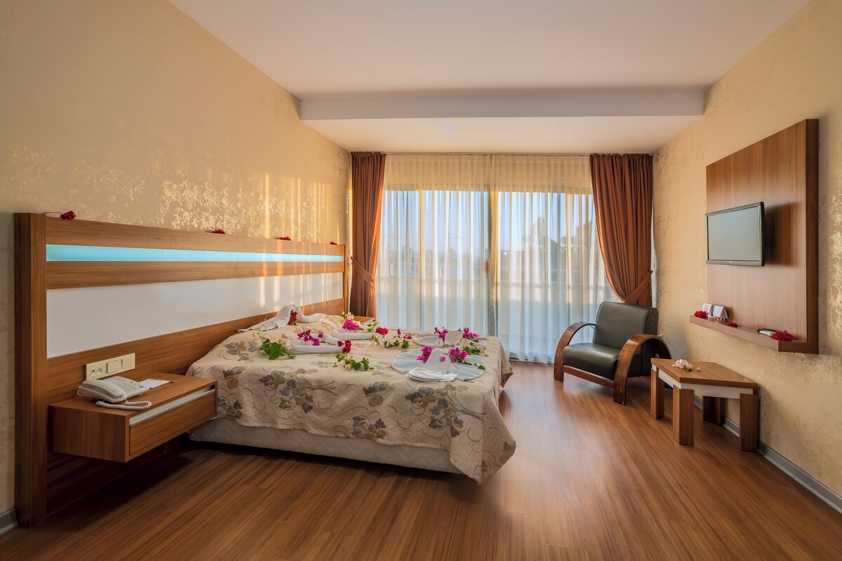 A hotel room with a bed, bedside table, chair, TV, and a small table with a flower.