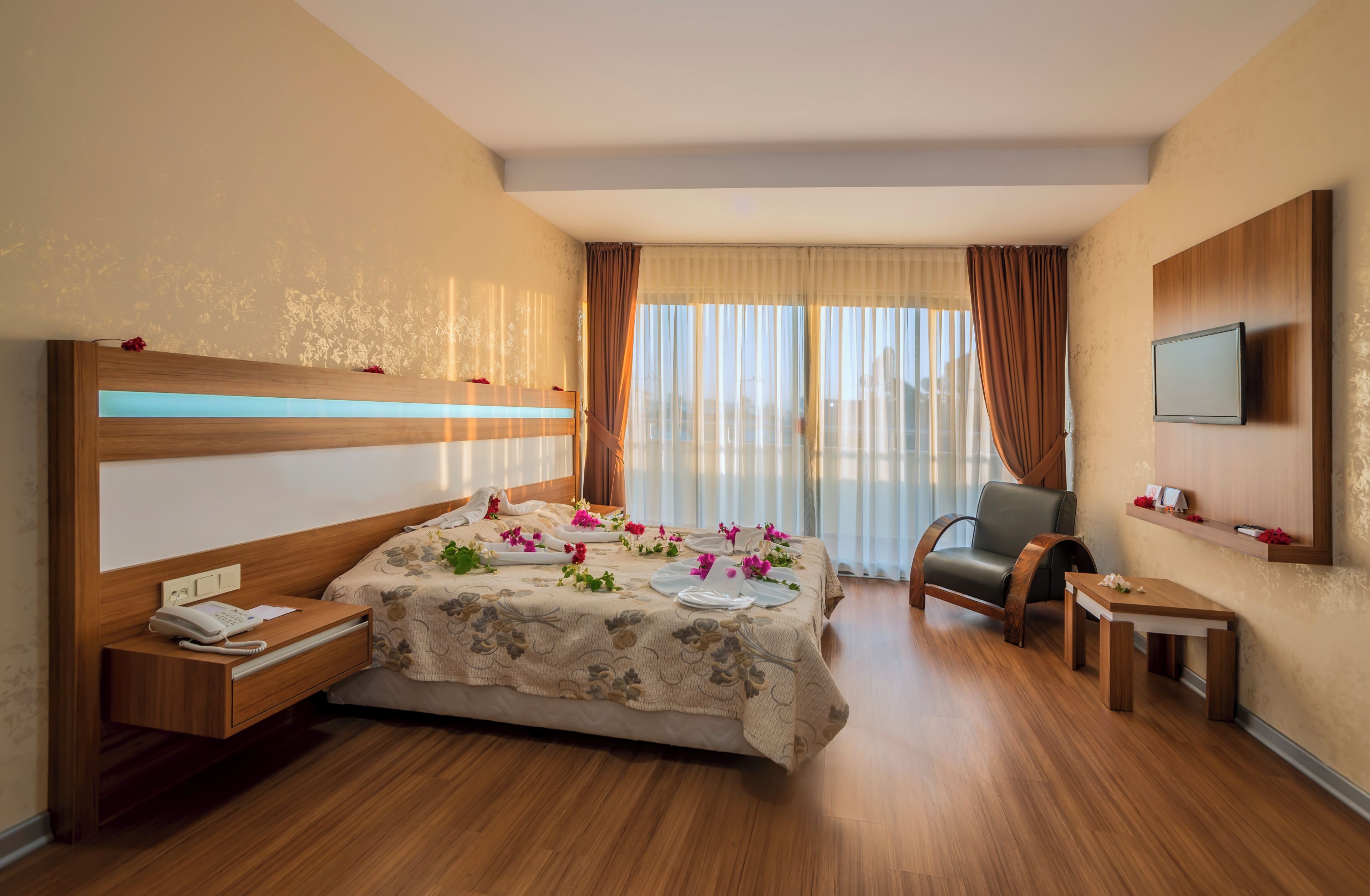 A hotel room with a bed, bedside table, chair, TV, and a small table with a flower.