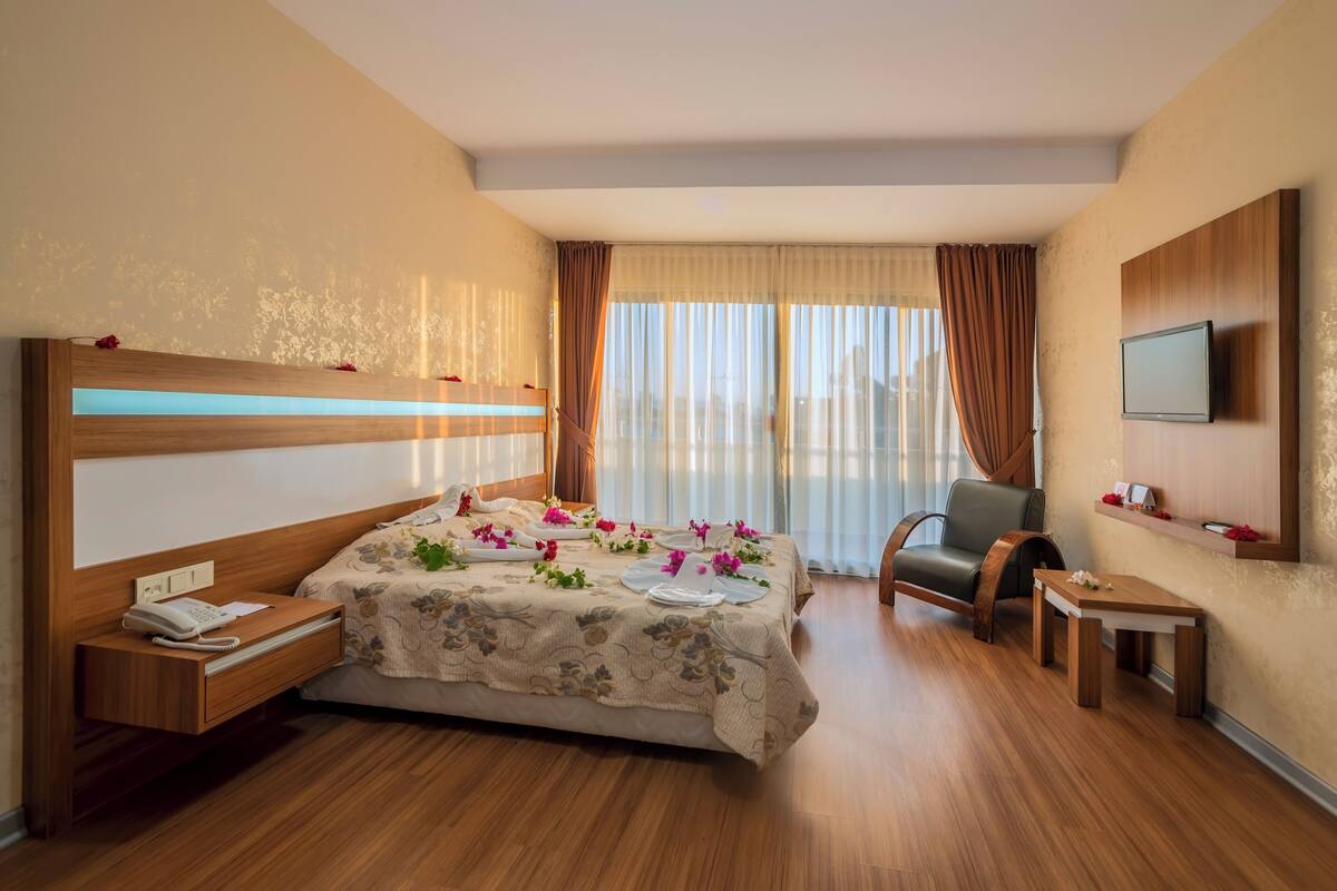 Een hotelkamer met een bed, nachtkastje, stoel, televisie en een kleintje tafeltje met een bloem.