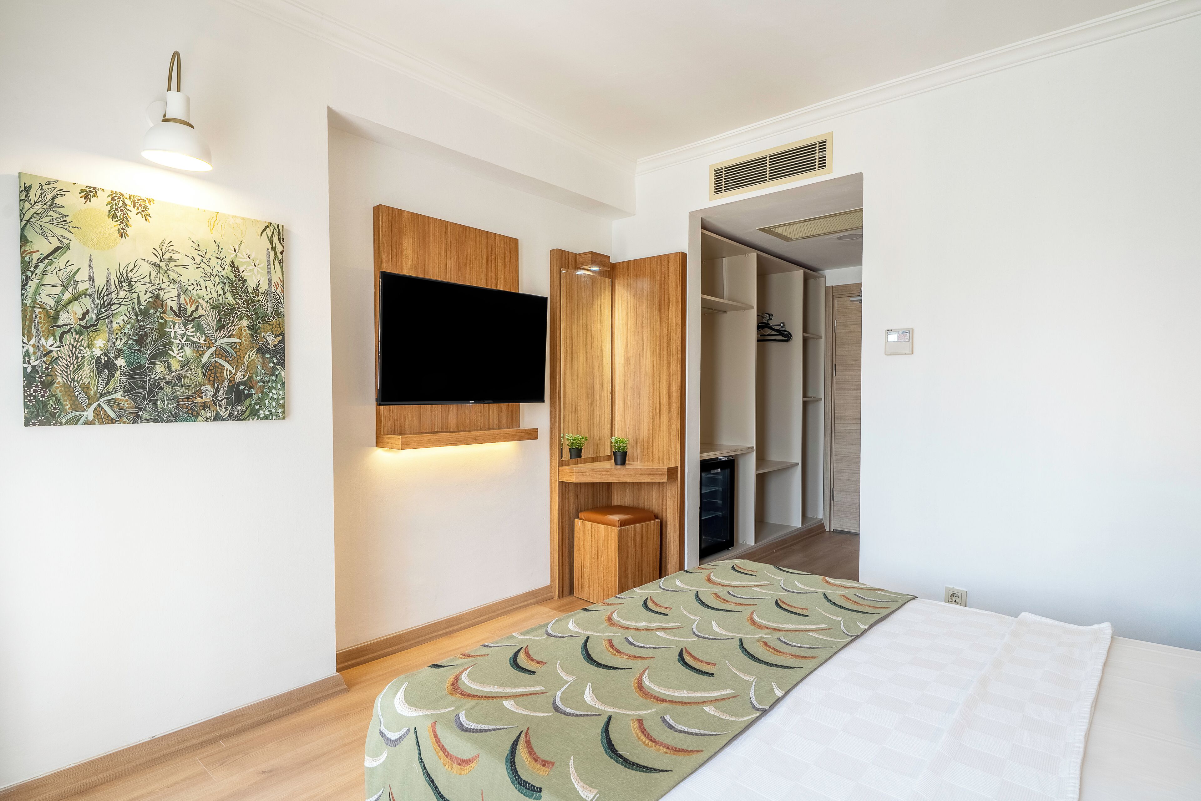 A bedroom with a bed, a mounted TV, a wooden wardrobe, a small shelf with plants, and a door leading to another room.