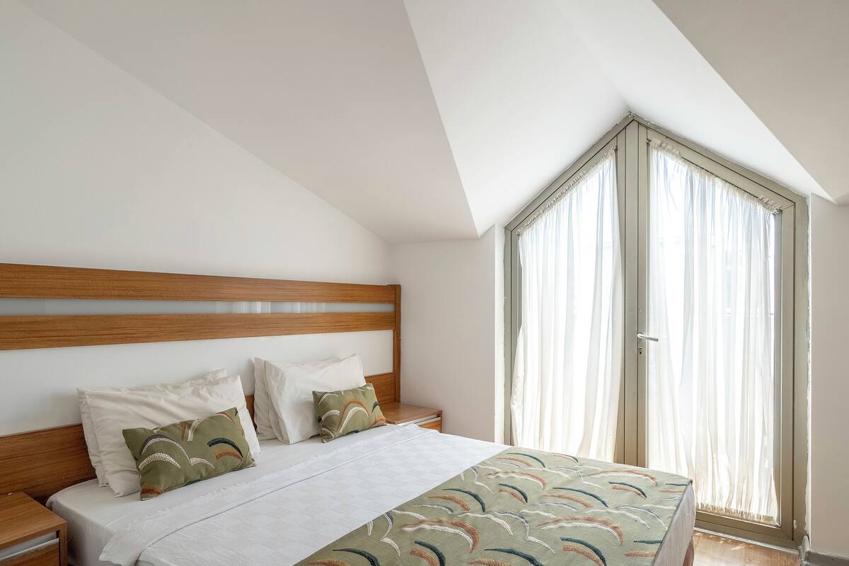 Een slaapkamer met een houten hoofdbord, een bed met beddengoed met een patroon, een nachtkastje en een groot raam met dunne gordijnen.