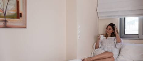 Uma pessoa sentada em uma cama em um quarto com um quadro na parede, uma janela com persiana e um interruptor de luz.