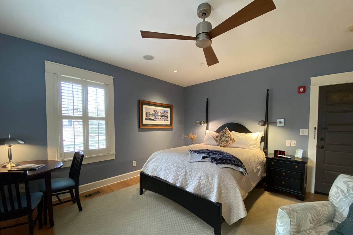 A bedroom with a bed, a desk, a chair, a ceiling fan, and a window.