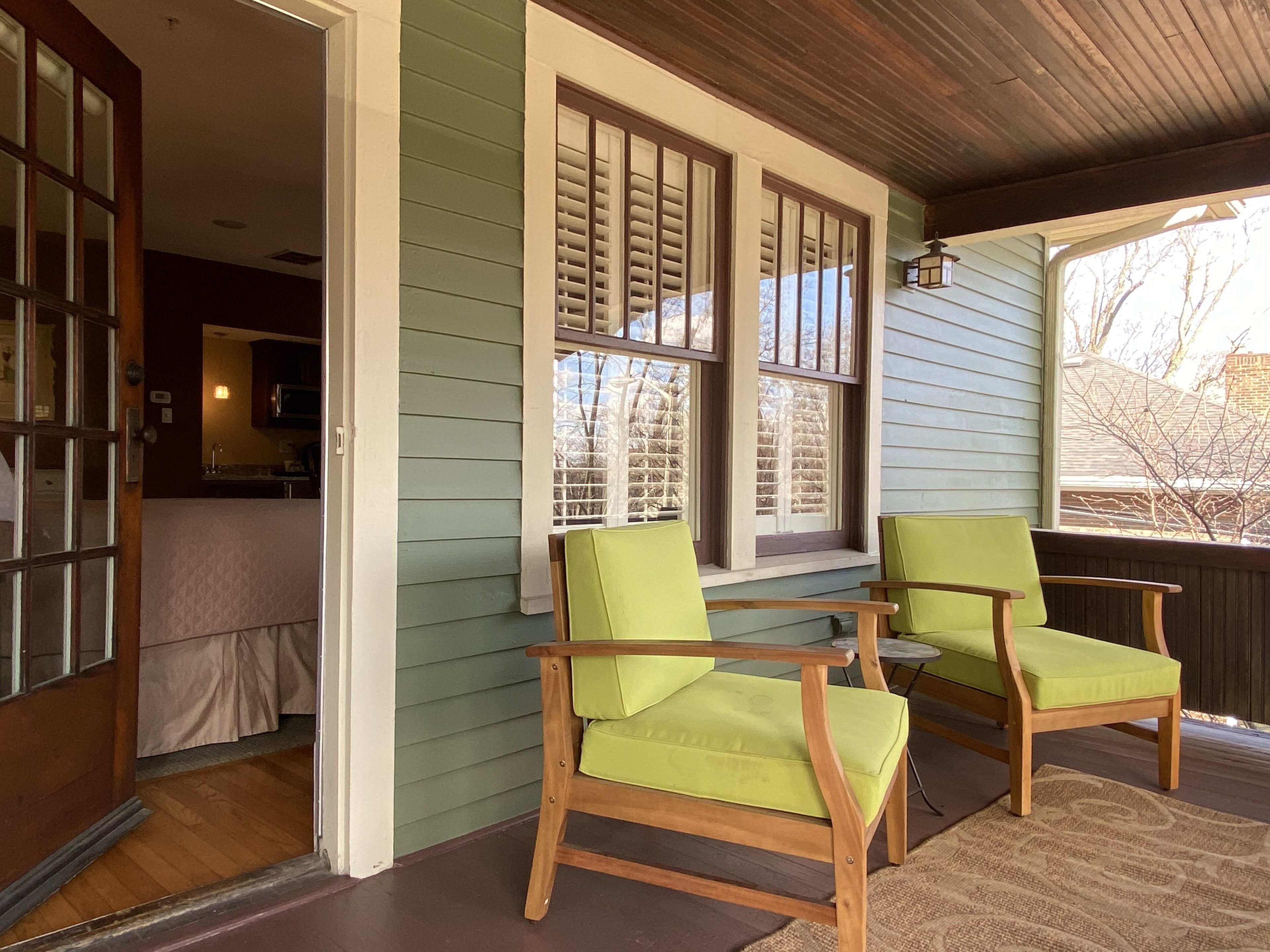 A porch with two wooden chairs and a table, a wooden ceiling, and a window with blinds.