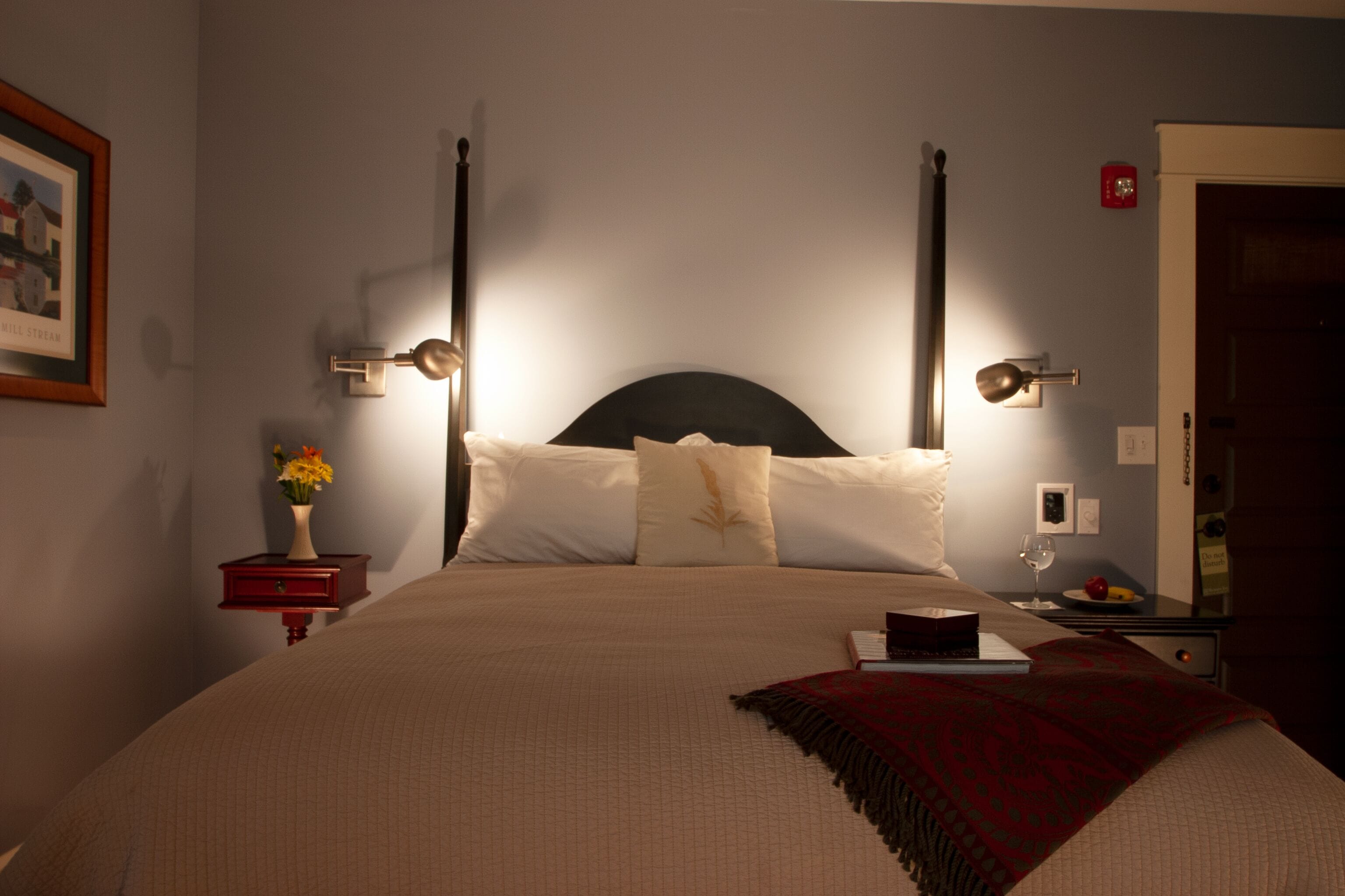 Ein ordentlich bezogenes Bett mit Kopfteil, zwei Nachttischlampen, einem kleinen Nachttisch mit Blumen in einer Vase und einem gerahmten Bild an der Wand.