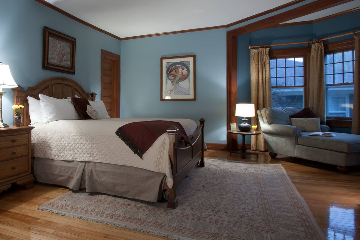 A bedroom with a large bed, a chair, a nightstand, a lamp, and a window with curtains.