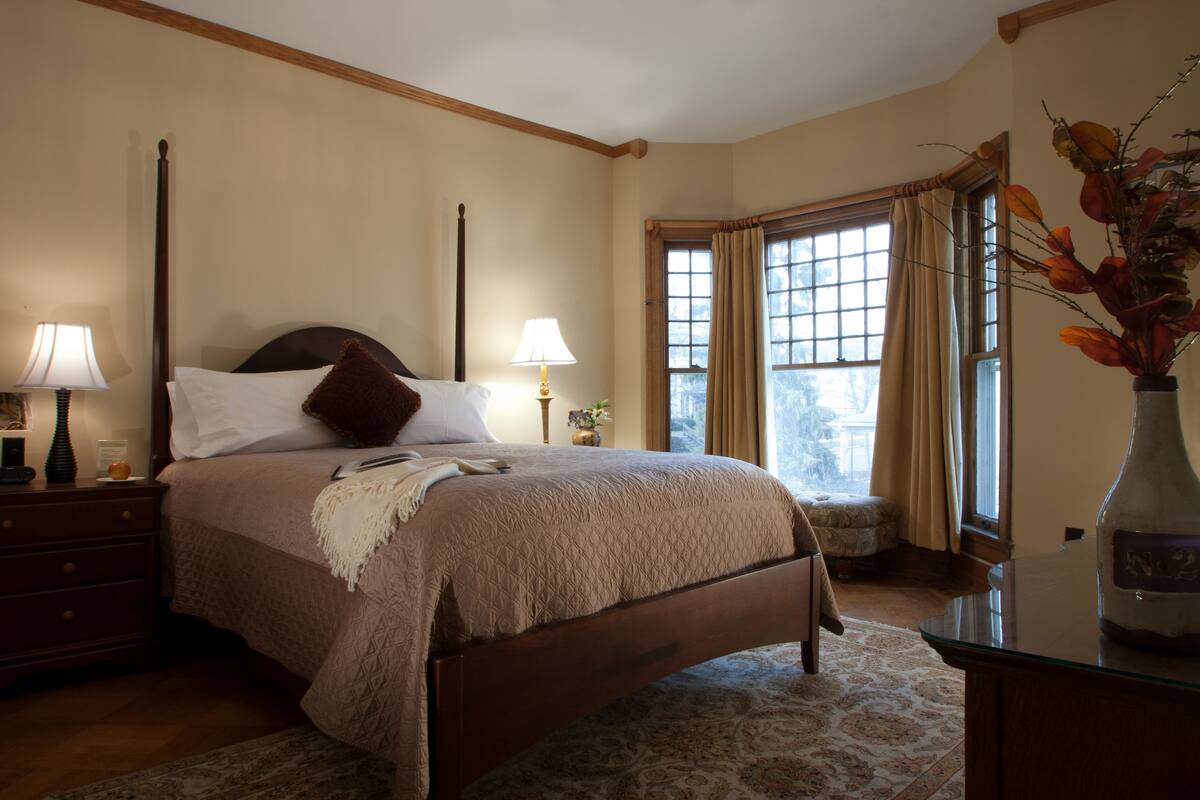 A bedroom with a four-poster bed, two bedside lamps, a nightstand, a vase with flowers, and a window with curtains.