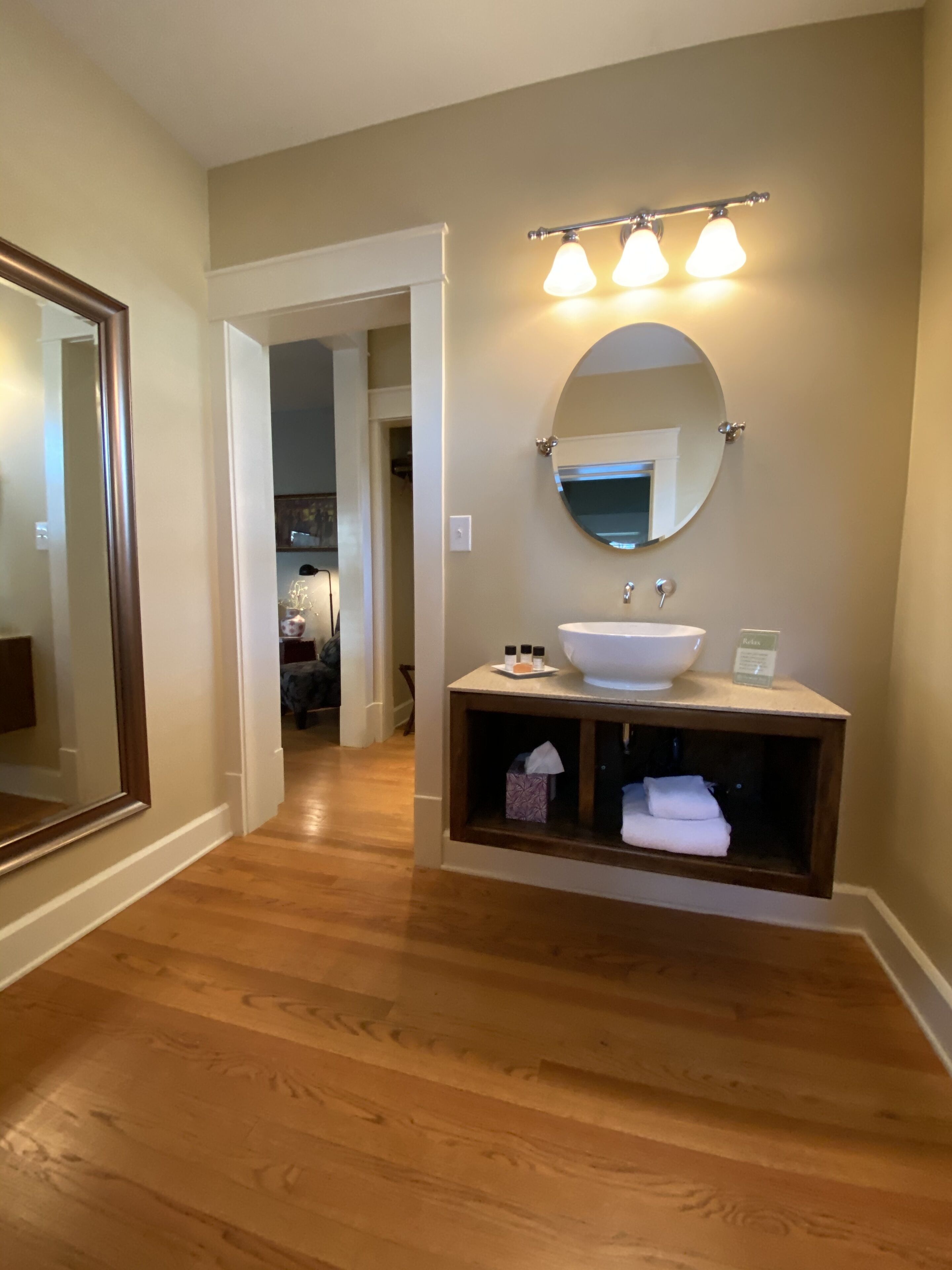 A bathroom with a wooden floor, a round mirror, a white sink, and a shelf with towels.