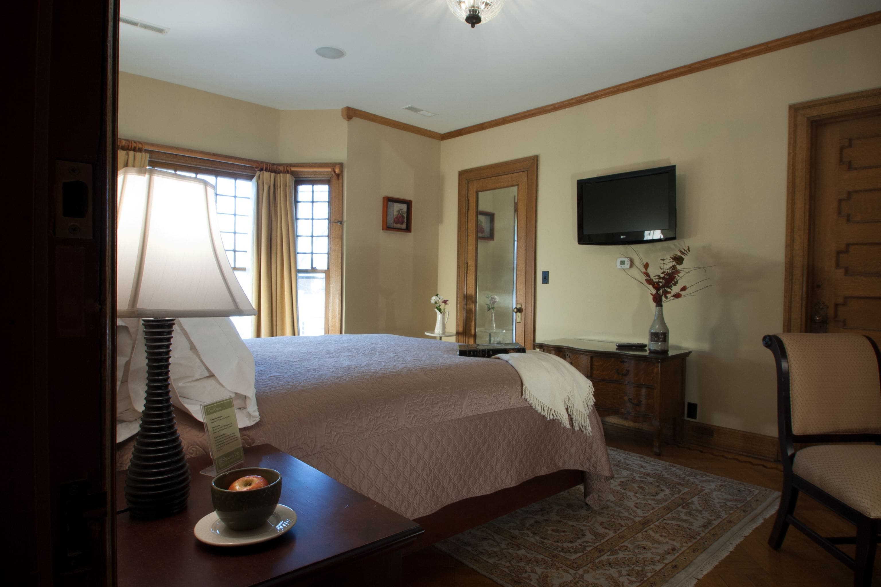 A hotel room with a bed, a television, a desk with a lamp, a vase with flowers, and a chair.