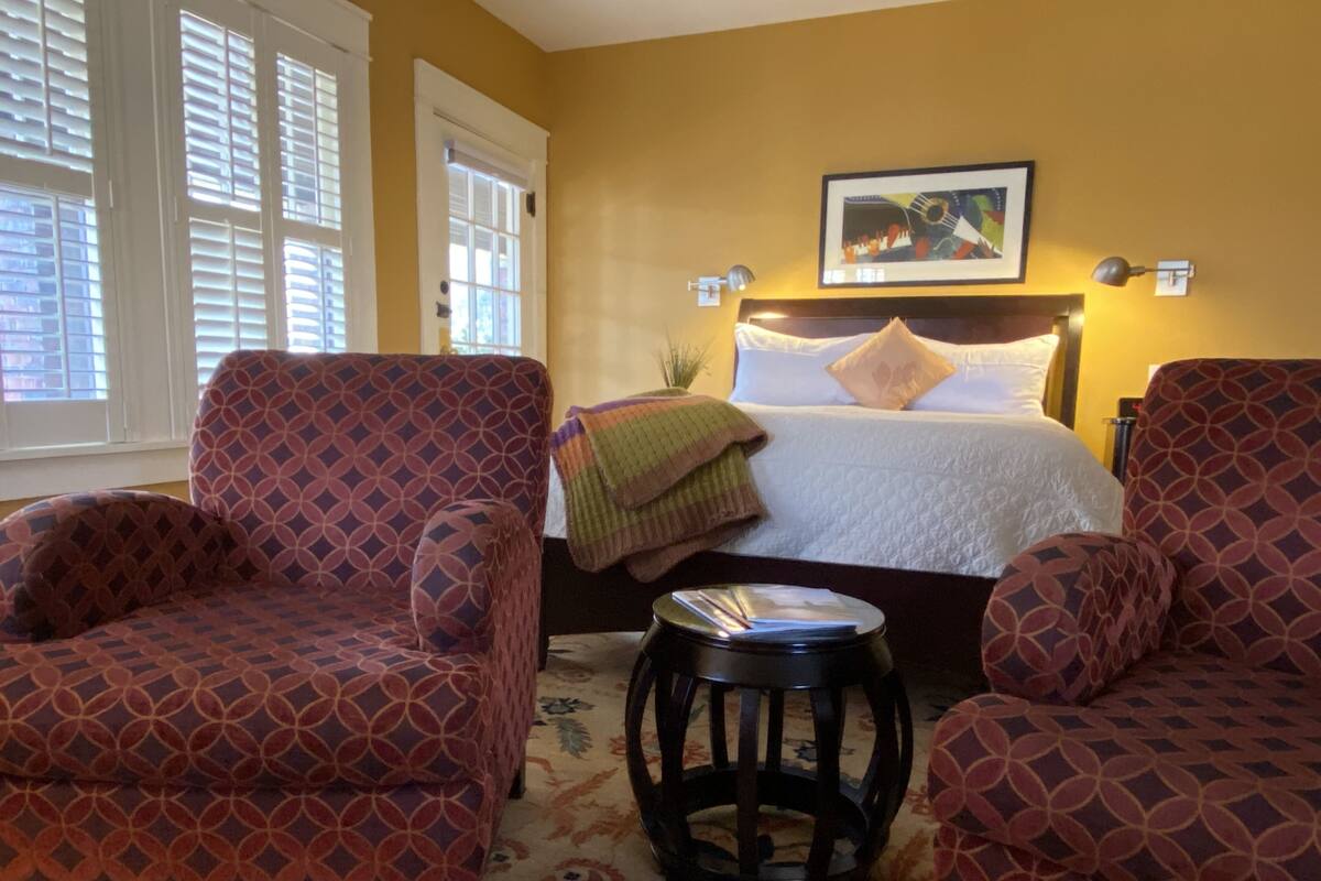 A hotel room with a bed, two patterned armchairs, a small round table, a painting on the wall, and a window with blinds.