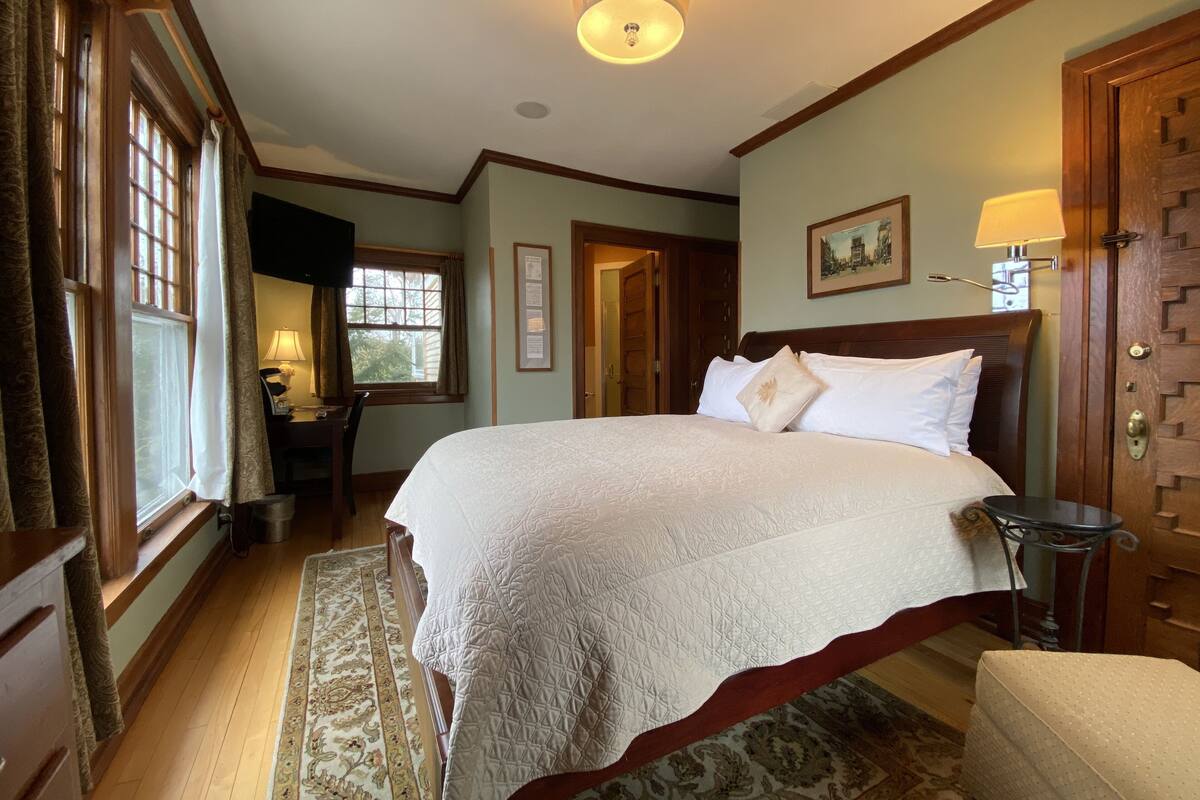 A well-lit bedroom with a large bed, a desk with a lamp, and a door leading to another room.