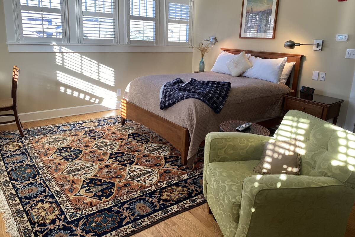 A bedroom with a bed, a nightstand, a lamp, a chair, and a window with blinds.