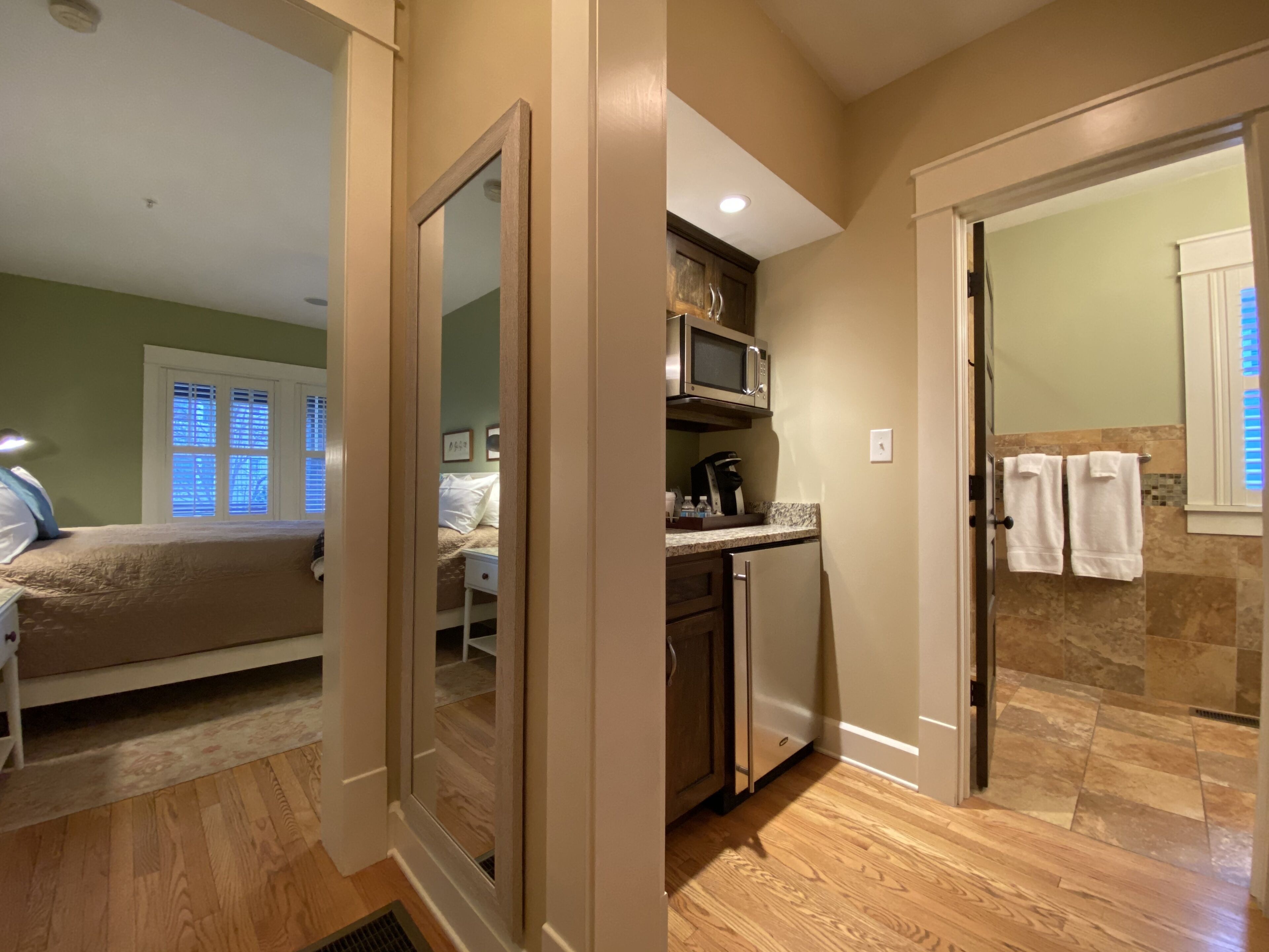 Une chambre à coucher comprenant un lit, une table de chevet et une commode. La salle de bain est équipée d’une douche, de toilettes et d’un lavabo. La chambre a un plancher en bois et une fenêtre avec des stores.