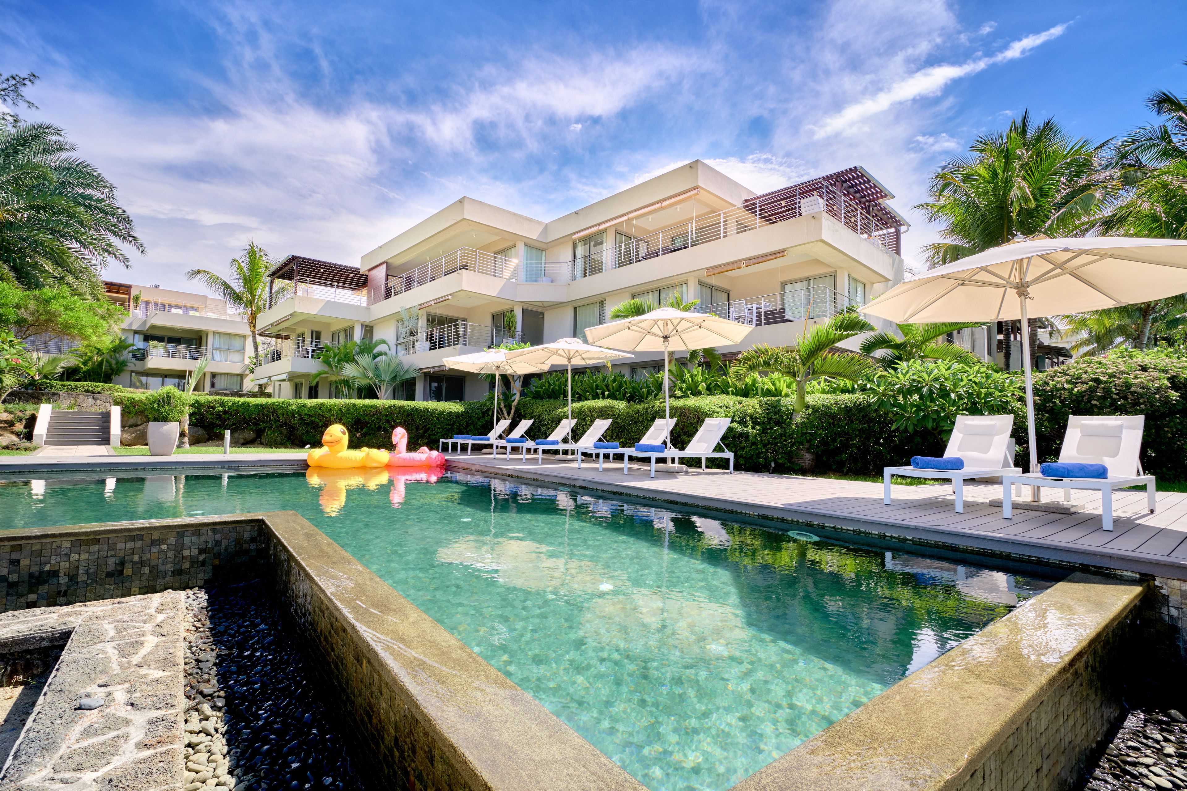 Une piscine entourée de chaises longues et de parasols, elle-même bordée d’un immeuble à plusieurs étages doté de balcons.
