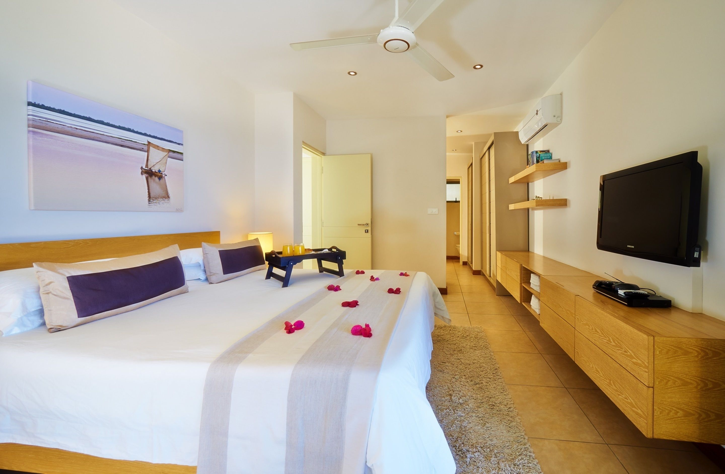 Habitación de hotel con una cama grande, ventilador de techo, televisor de pantalla plana y mueble de televisión de madera.