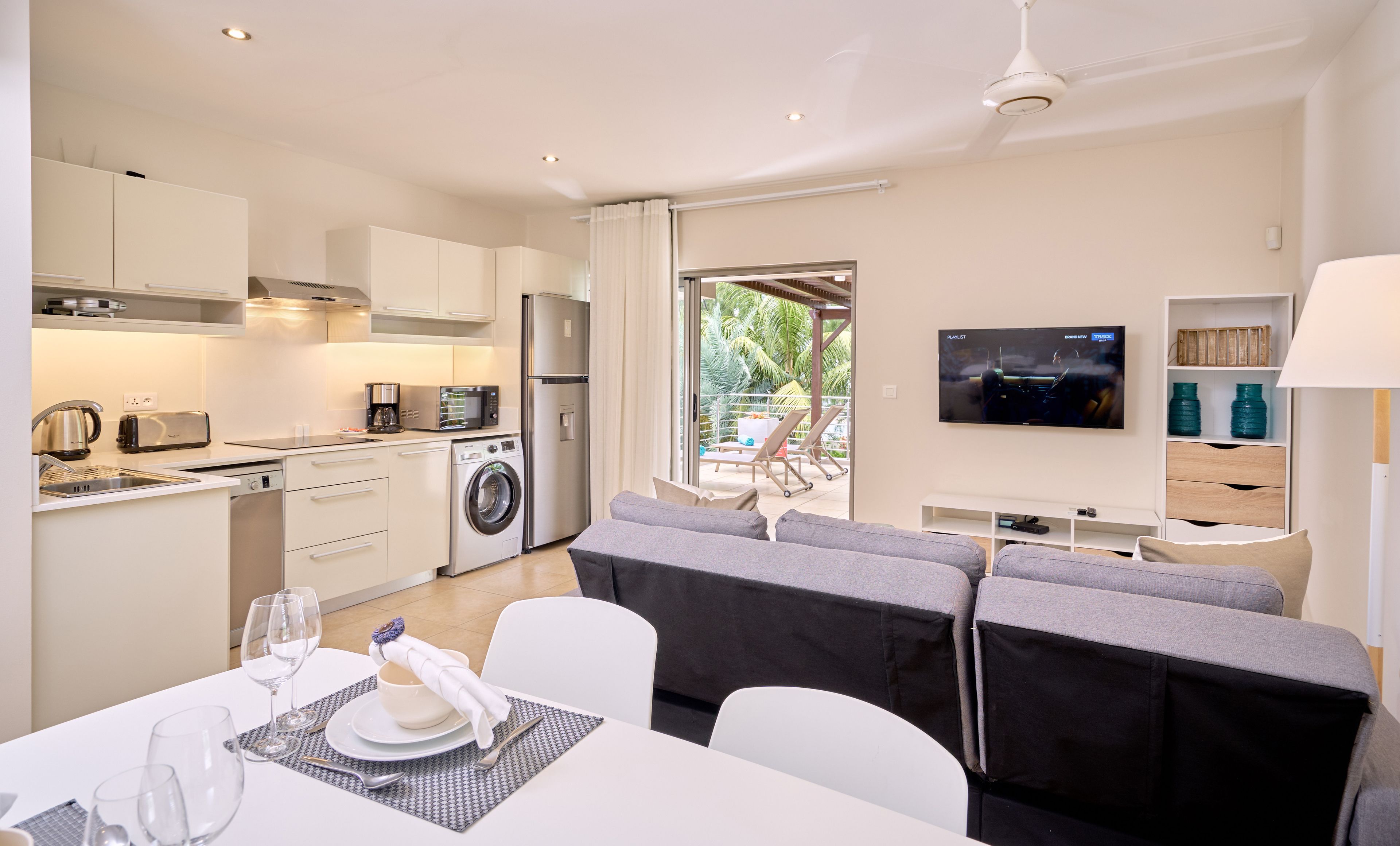Cocina moderna con lavadora integrada, comedor con mesa lista para dos personas y televisor montado en la pared.