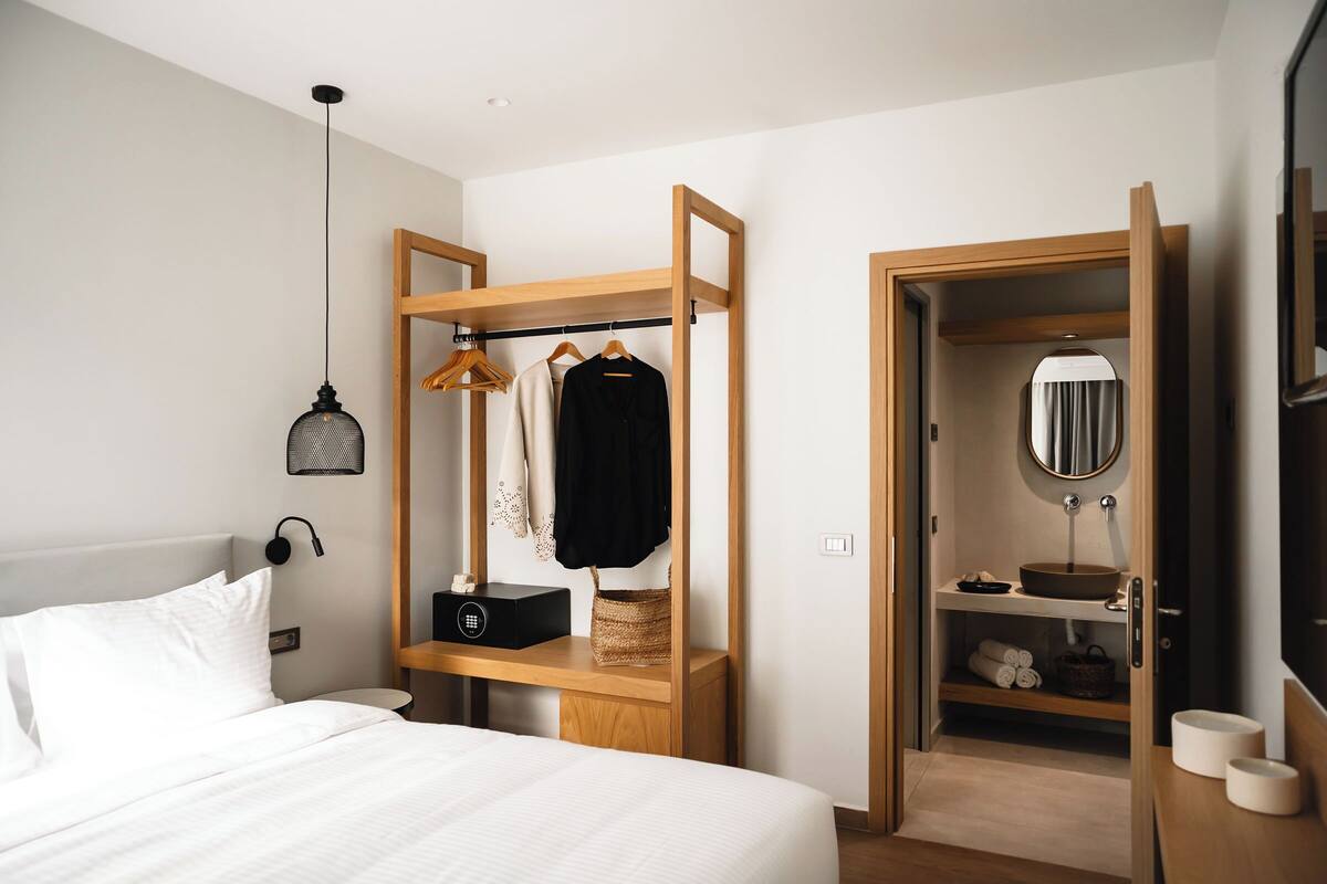 Un dormitorio con cama, mesita de noche de madera, lámpara colgante, armario y baño con lavabo y espejo.