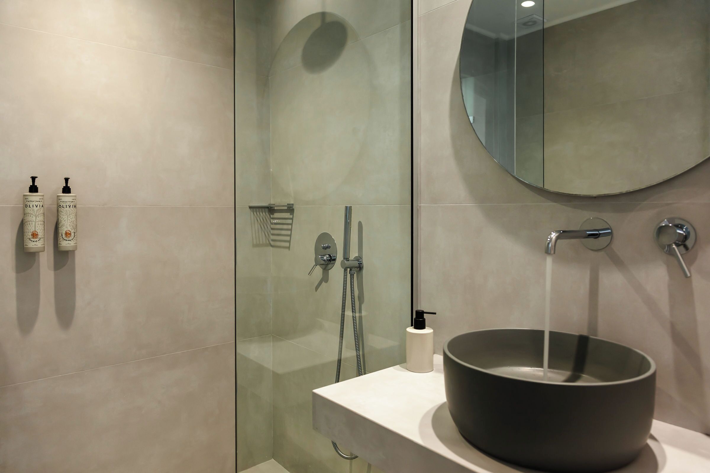 Un baño moderno con ducha de vidrio, espejo redondo y lavabo negro sobre encimera blanca.