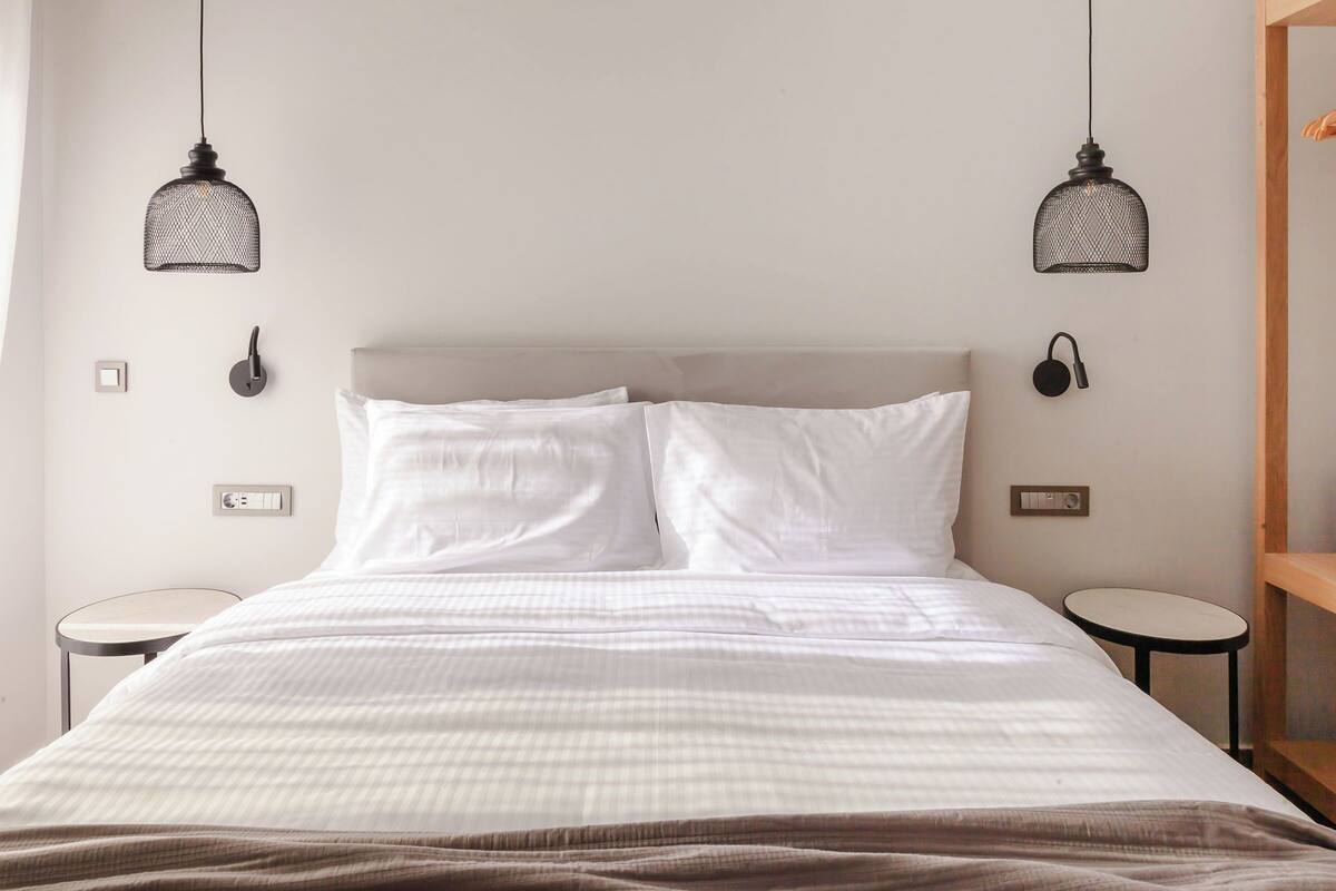 Una cama bien hecha con sábanas blancas, dos mesitas de noche y dos lámparas colgantes.