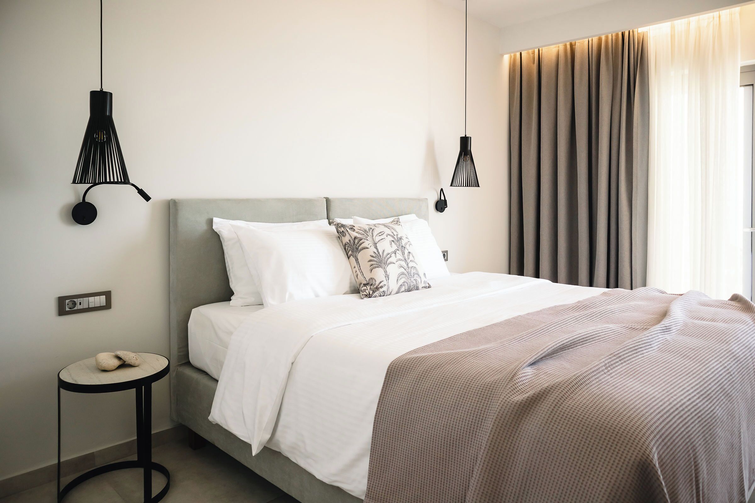 A neatly made bed with a grey headboard, white bedding, and a patterned pillow. There is a small round table with a cushion, a wall-mounted light fixture, and a window with curtains.