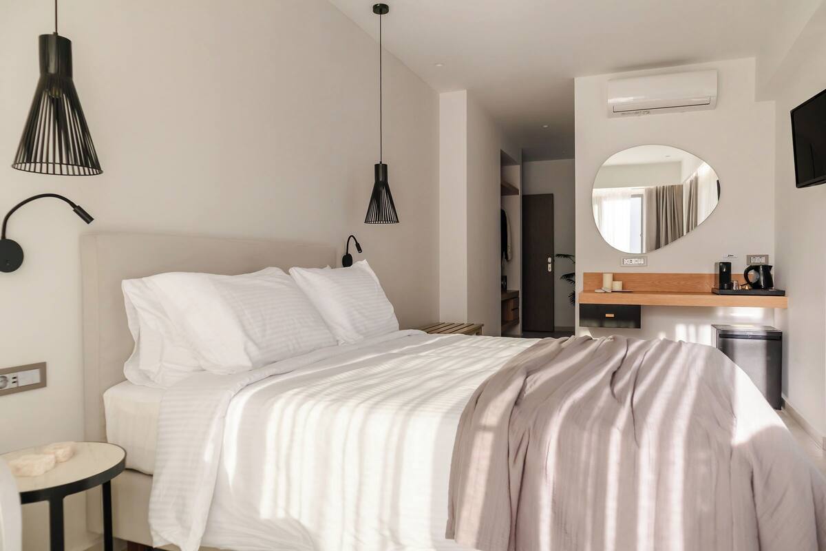 A modern hotel room with a bed, bedside table, lamp, mirror, and a small kitchenette area.