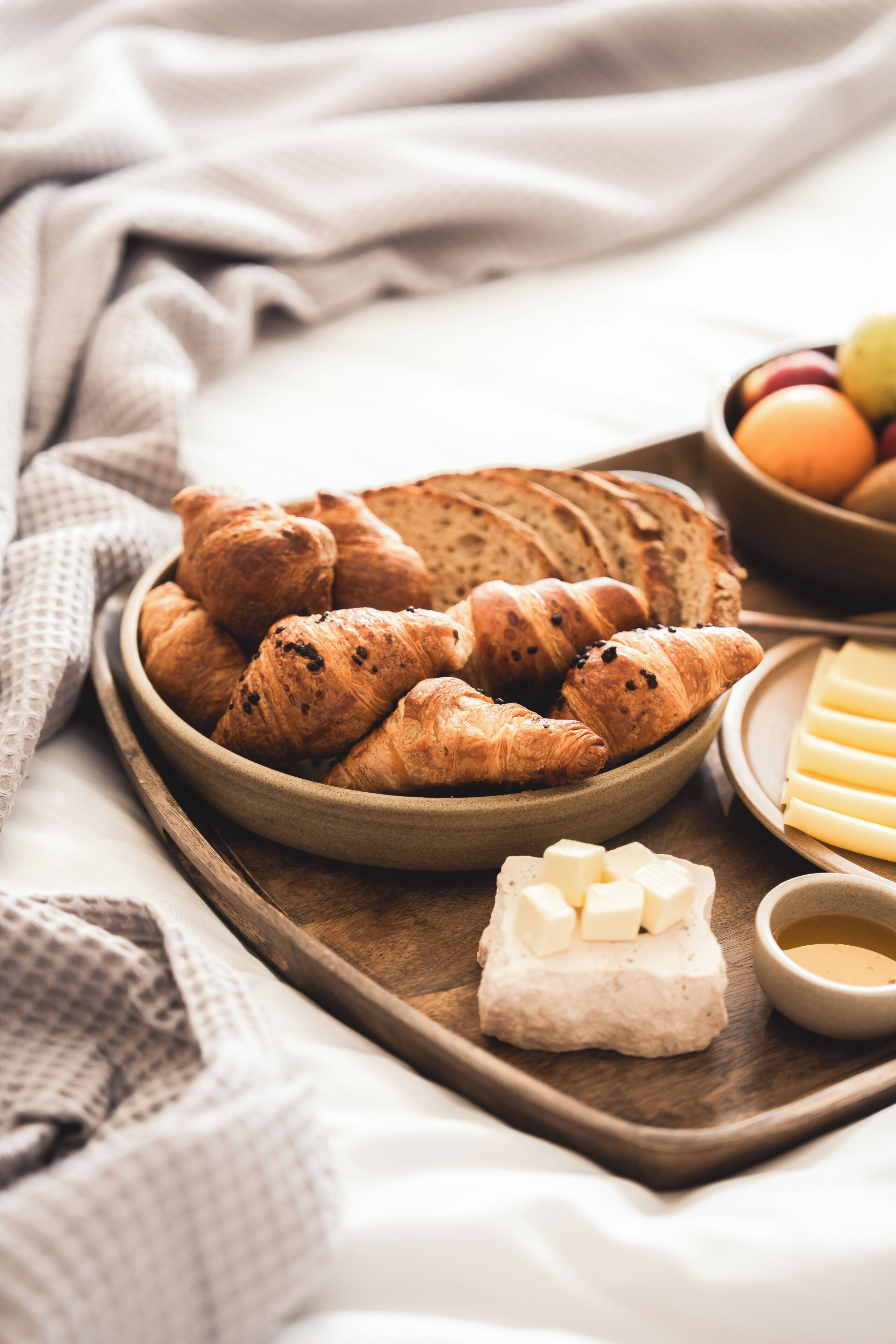 Uma bandeja de madeira com croissants, queijo e uma tigela de frutas.
