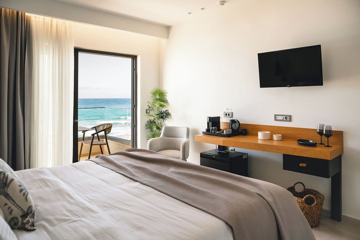 Una habitación de hotel moderna con una cama, un escritorio con televisor de pantalla plana, una silla, una planta pequeña y vista al océano.