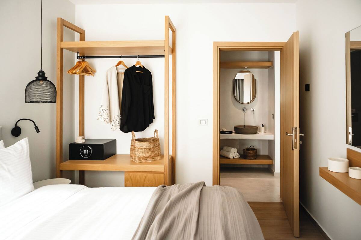 A bedroom with a bed, a wooden shelf with clothes and a basket, a wardrobe, and a bathroom with a mirror and sink.