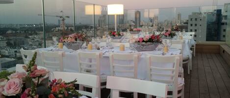 Un área de comedor en la azotea con mesas dispuestas para una comida, con vista a un paisaje urbano.