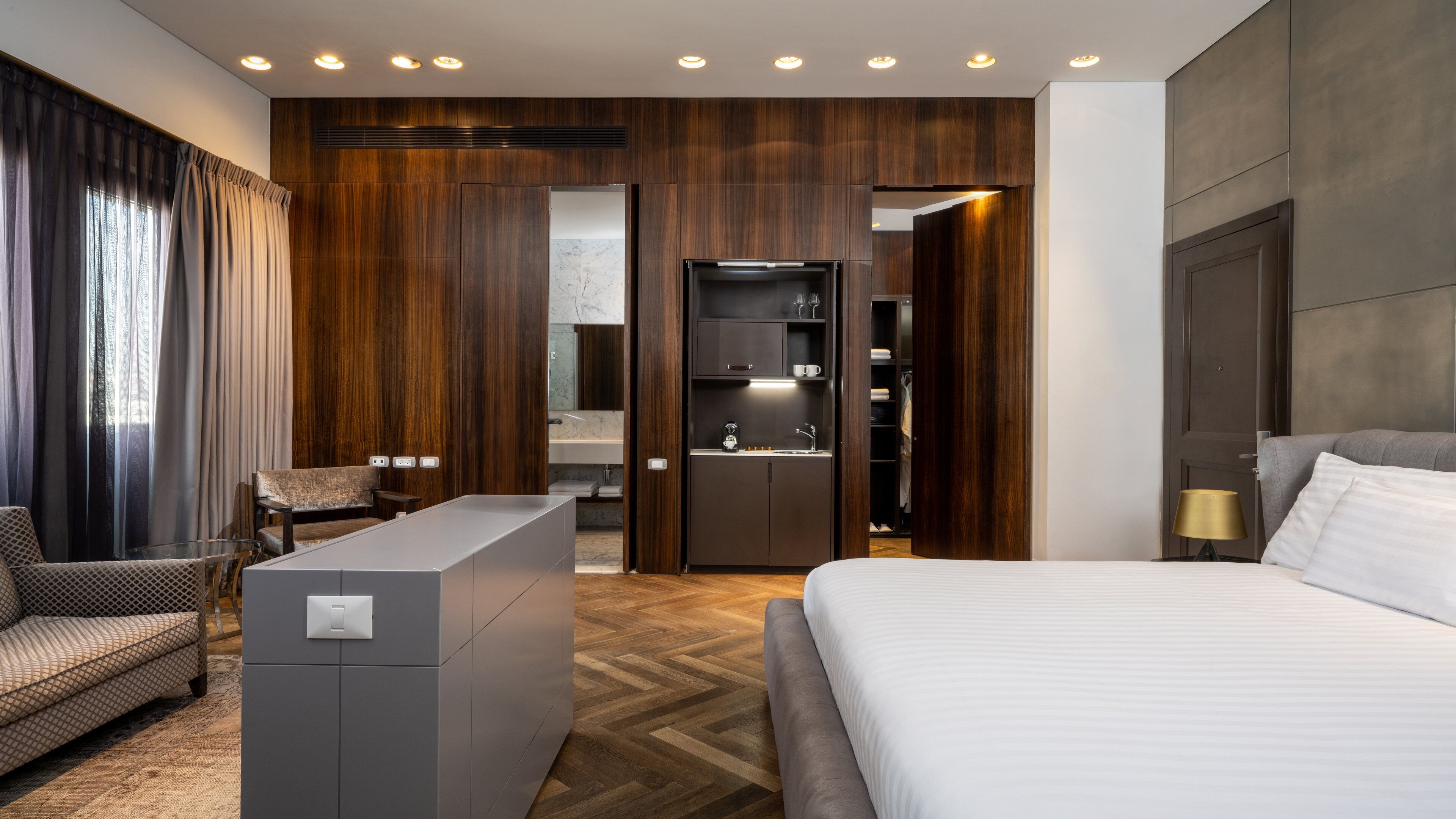 A hotel room with a bed, a seating area, a minibar, and a wardrobe.