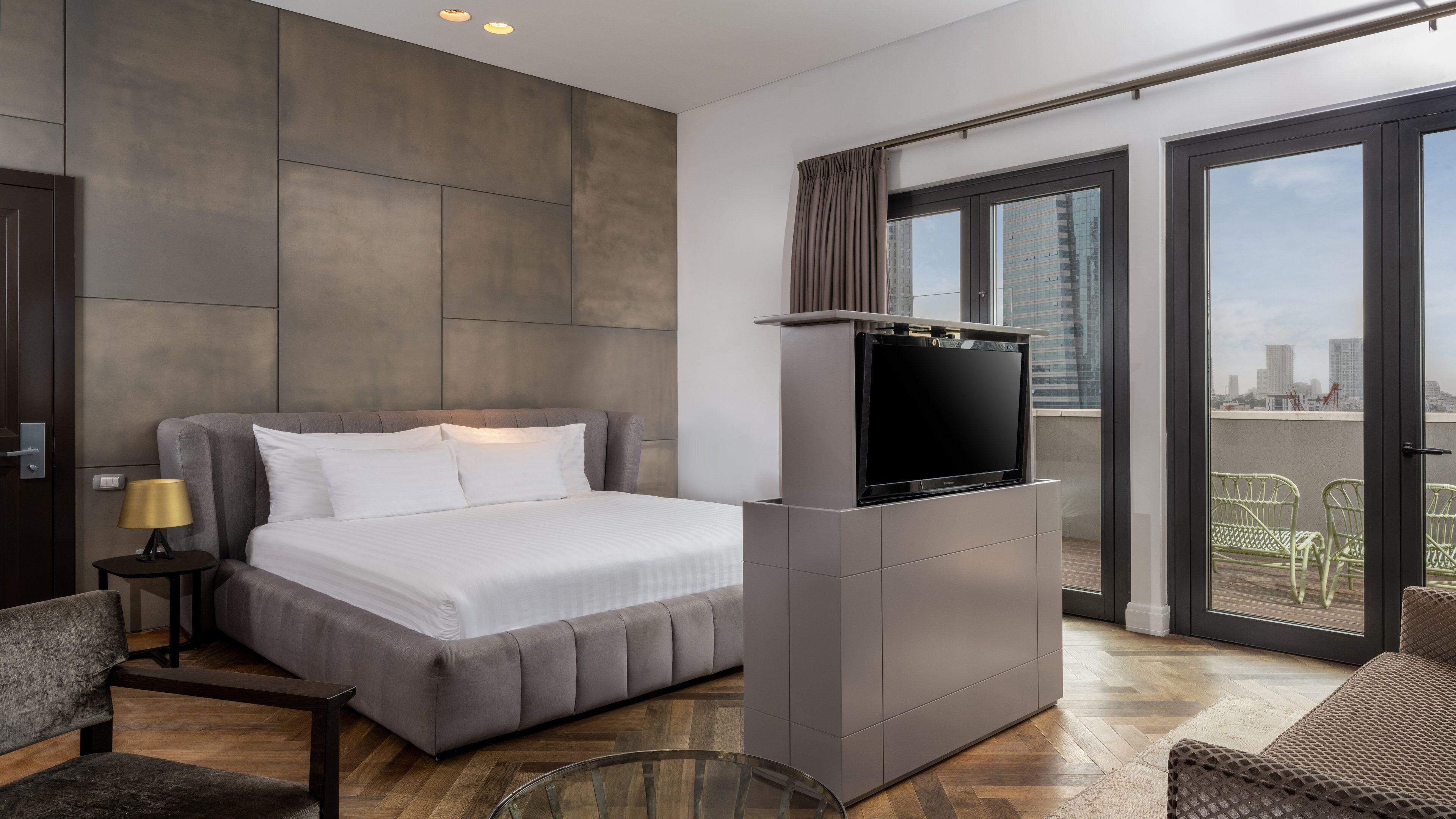 A modern hotel room with a large bed, a flat-screen TV, a balcony with a view of the city, and a small seating area.