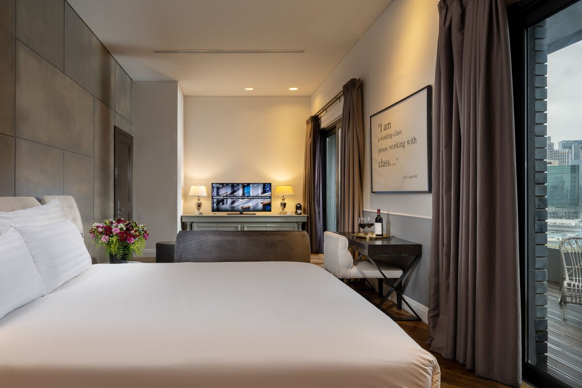 Ein modernes Hotelzimmer mit einem großen Bett, einem Schreibtisch mit Computer, einem Fernseher und Blick auf die Stadt durch große Fenster.