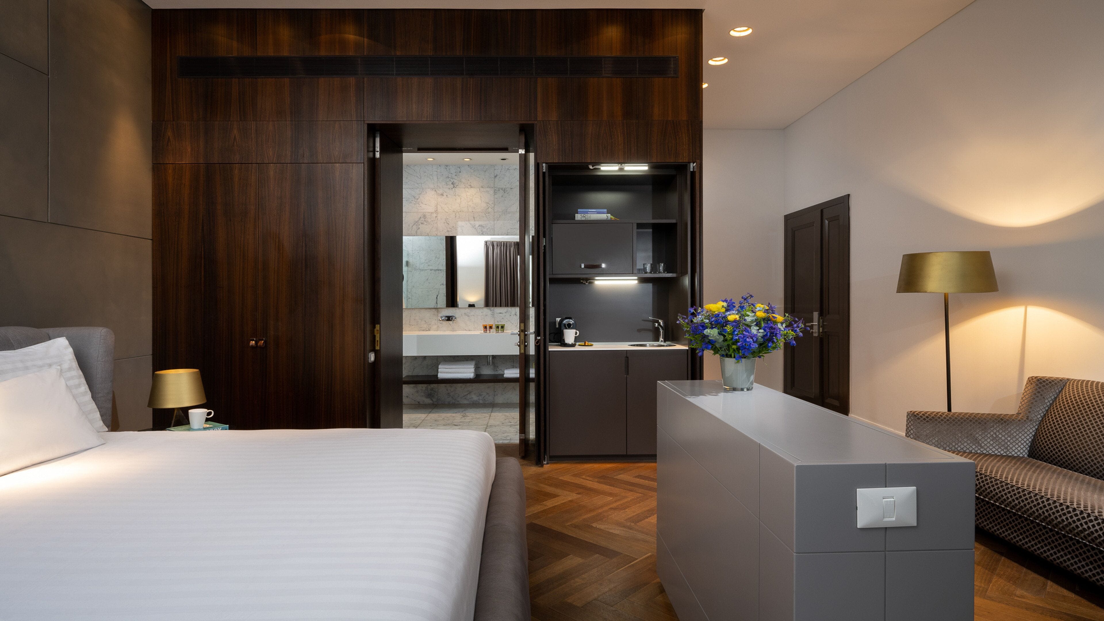 Een moderne hotelkamer met een groot bed, een bureau met een bloemenvaas, een zithoek en een aparte badkamer met wastafel en spiegel.