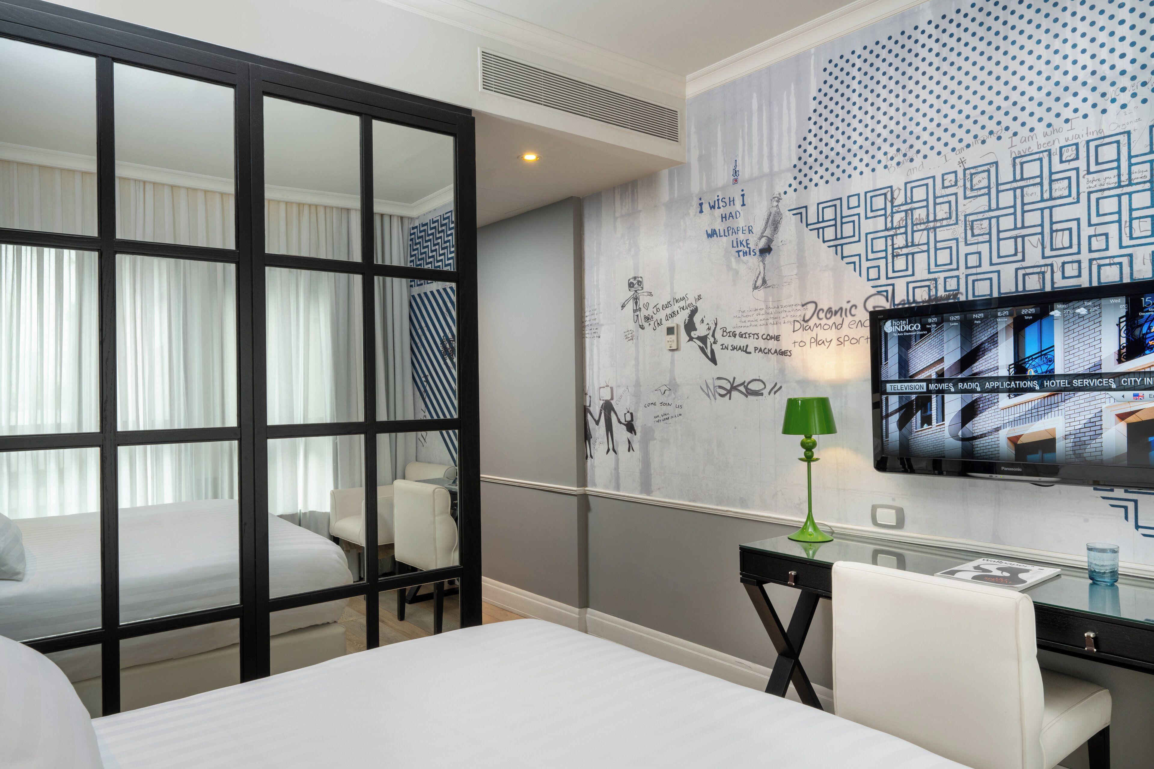A hotel room with a bed, a desk with a lamp, a TV on the wall, and a decorative wall with patterns and text.