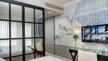 A hotel room with a bed, a desk with a lamp, a TV on the wall, and a decorative wall with patterns and text.