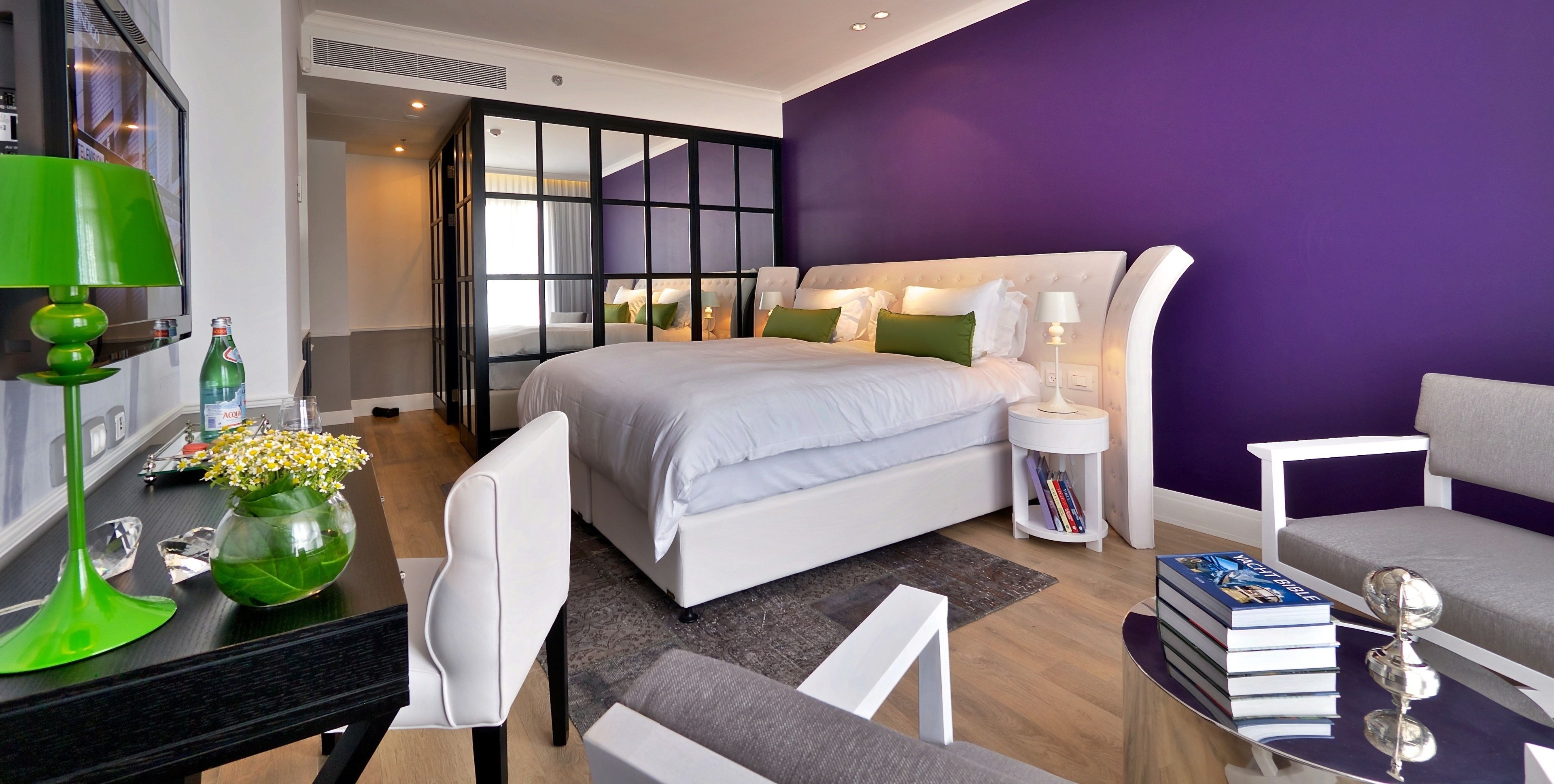 Une chambre d’hôtel moderne avec un grand lit, un bureau avec une lampe, une chaise, une bibliothèque et un mur d’accent violet.