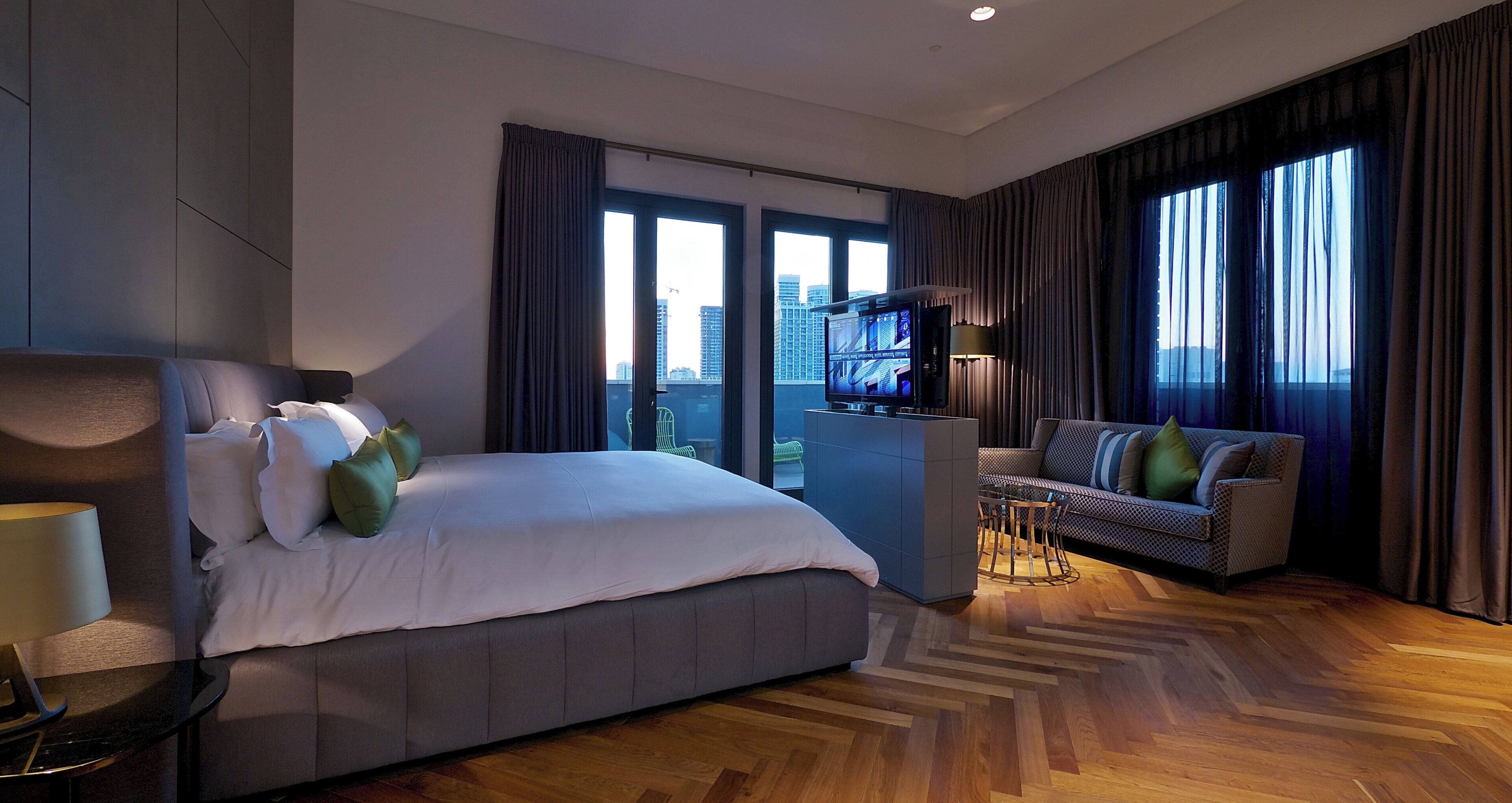 Une chambre d’hôtel moderne dotée d’un grand lit, d’un coin salon avec un canapé et des fauteuils, d’une télévision et offrant une vue sur le paysage urbain grâce à de grandes fenêtres.