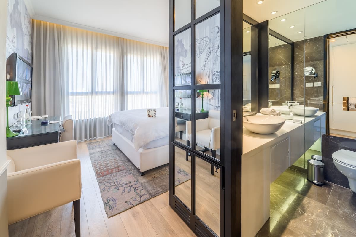 Een moderne hotelkamer met een groot bed, een zithoek met een bank en fauteuil, een aparte badkamer met een grote spiegel, en een televisie in de hoek.