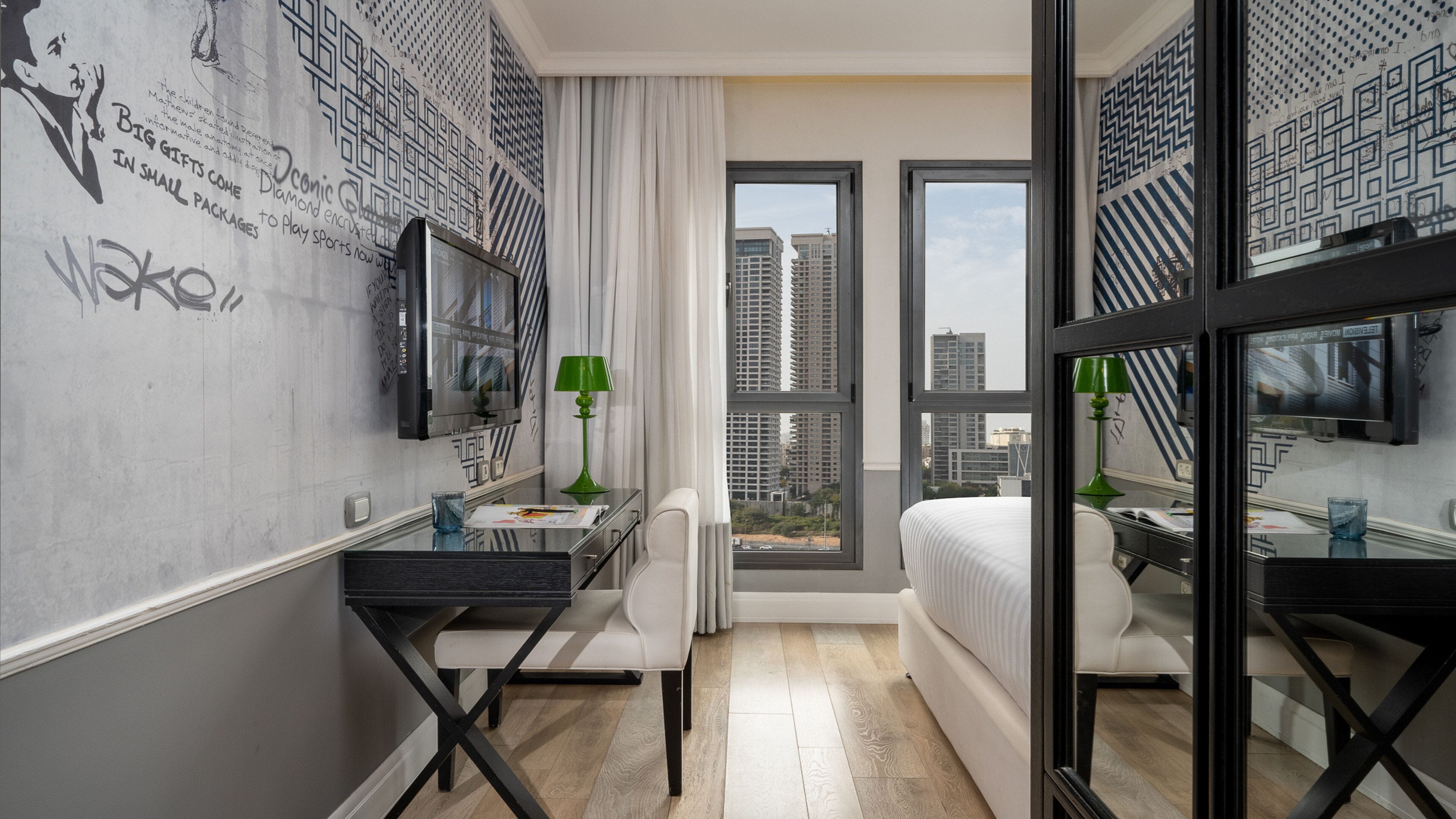 A modern hotel room with a large bed, a desk with a chair, a television, and a window with a city view.