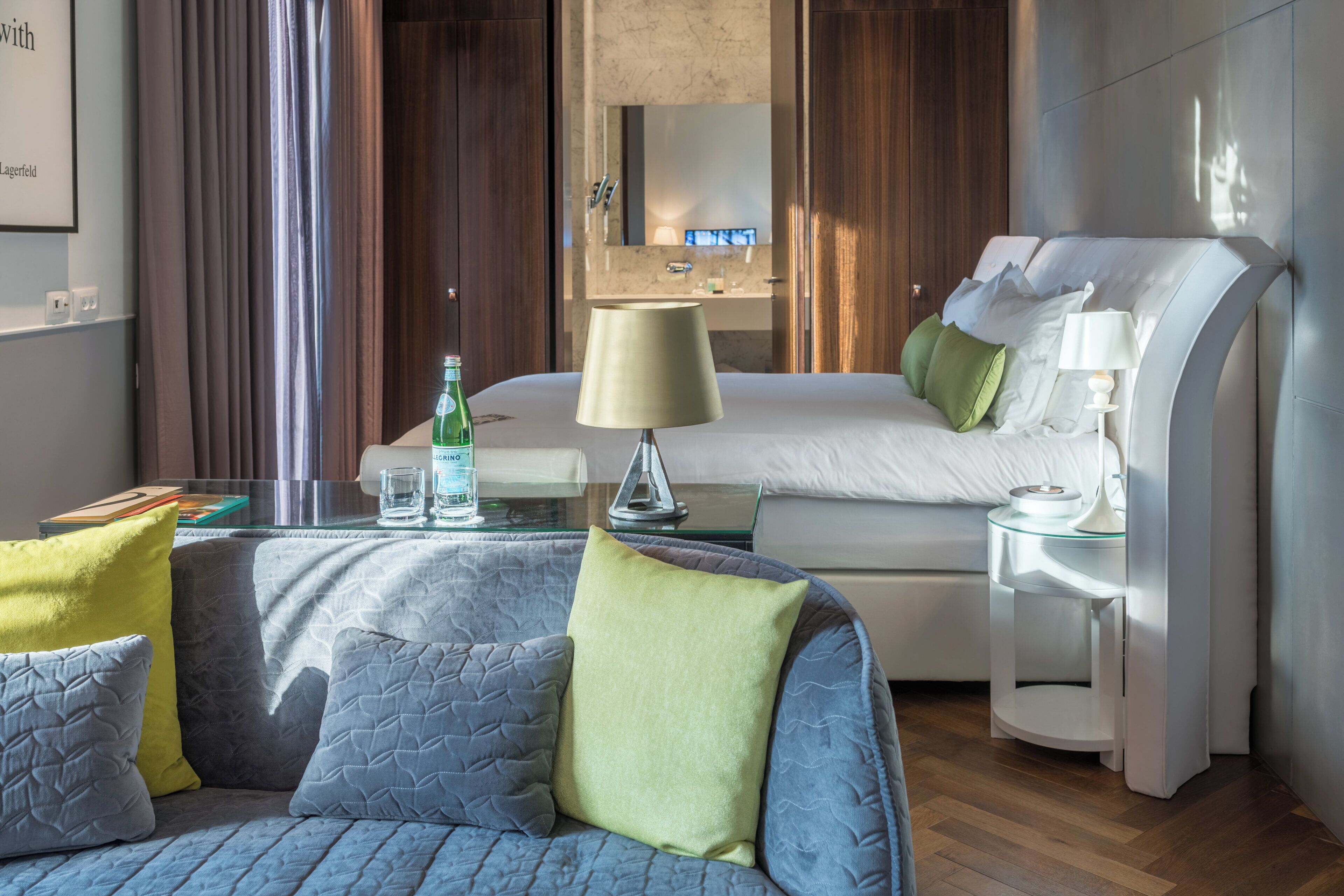 Una habitación de hotel con cama, sofá, mesita de noche, lámpara y vaso de agua.