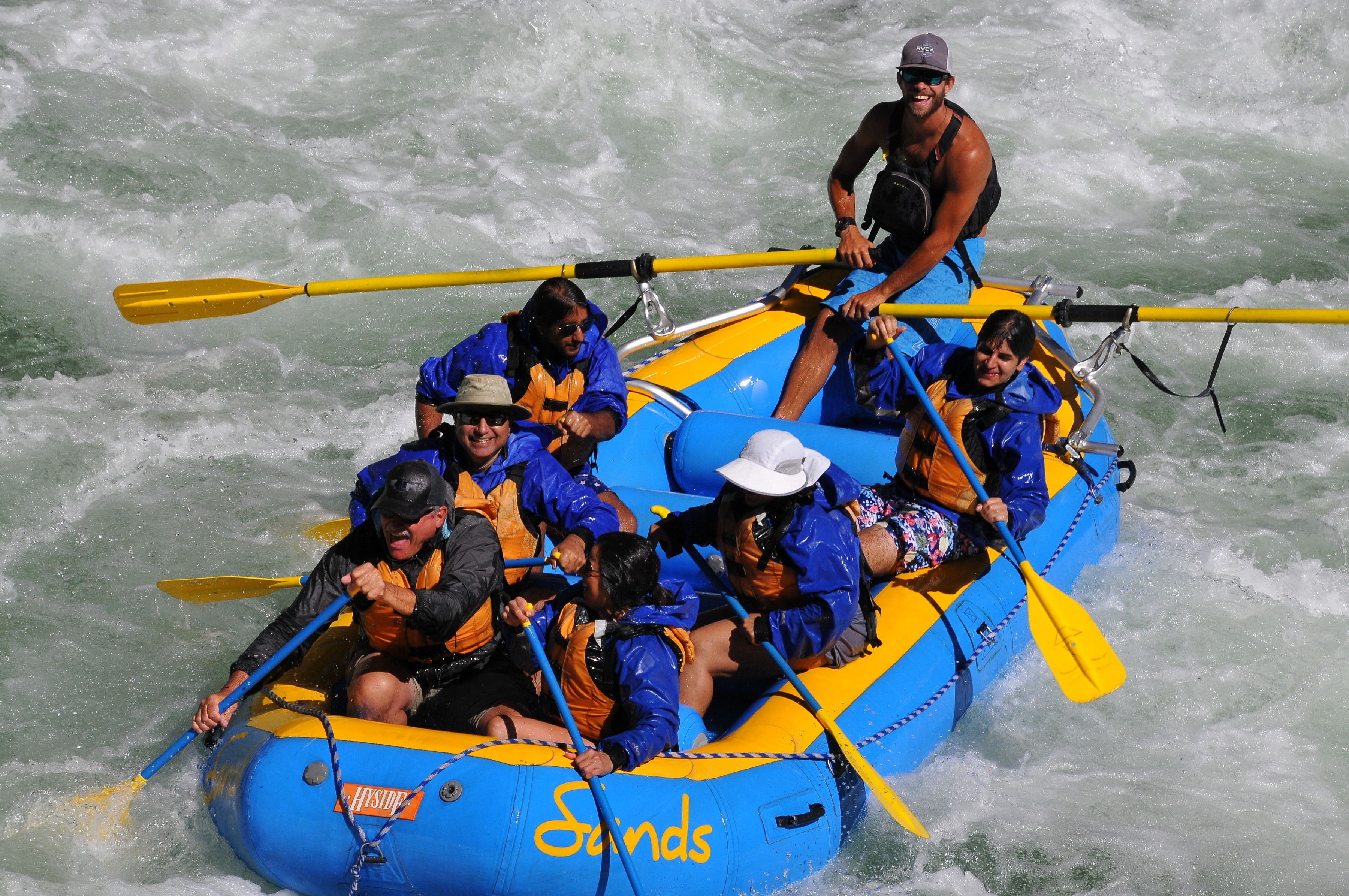 A group of people in a blue and yellow raft with paddles, navigating through white water.