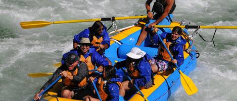 Un groupe de personnes à bord d’un radeau bleu et jaune, équipées de pagaies, navigue dans des eaux tumultueuses.