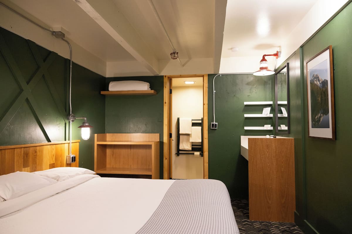 A hotel room with a bed, a wooden headboard, a wooden shelf, a bathroom with a sink and mirror, and a framed picture on the wall.