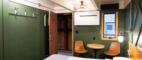 Une pièce comprenant un lit, deux chaises, une petite table, une fenêtre et un appareil de climatisation.