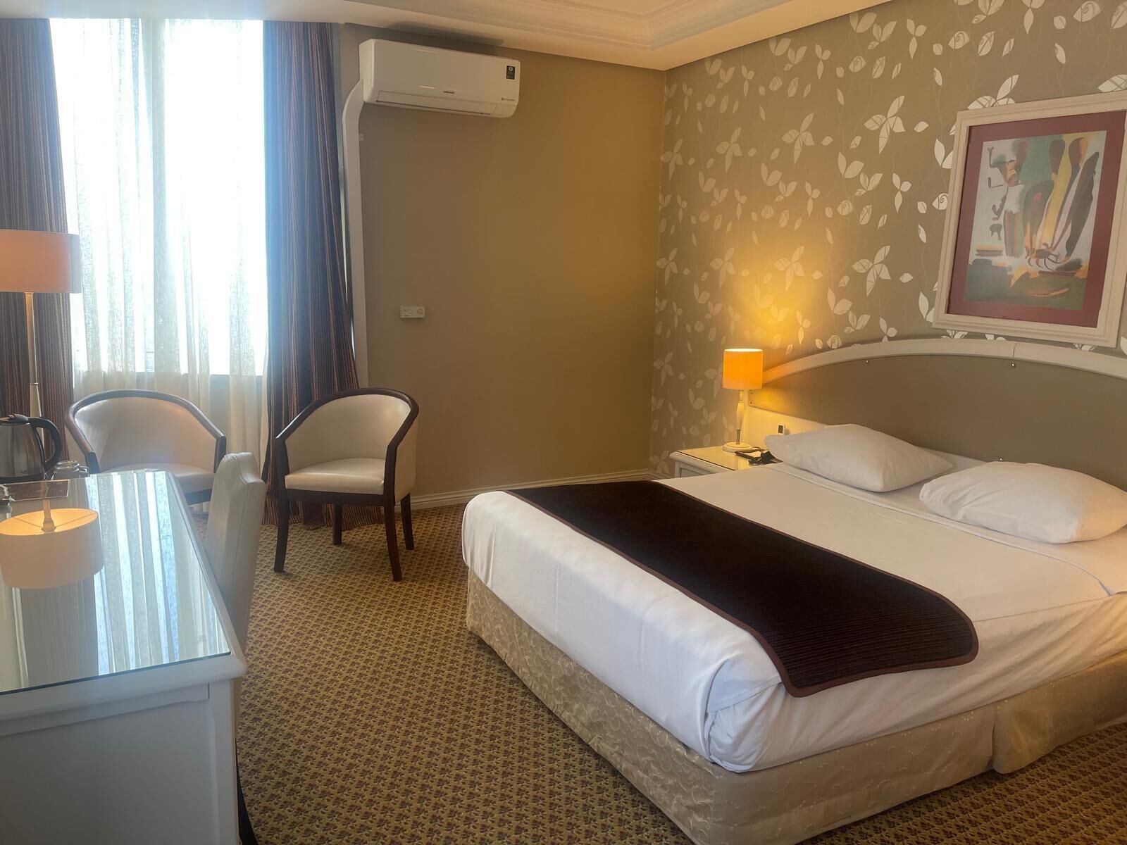 A hotel room with a bed, two chairs, a desk, a lamp, and an air conditioning unit.