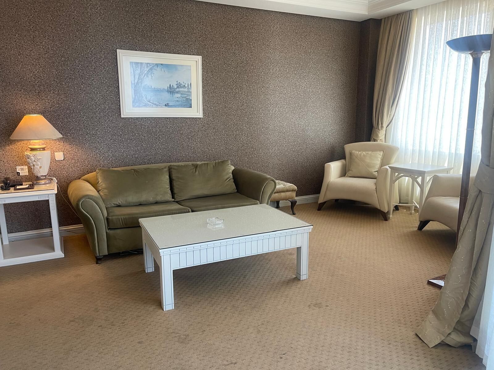 A hotel room with a beige sofa, a white coffee table, a beige armchair, a small white table, and a lamp.