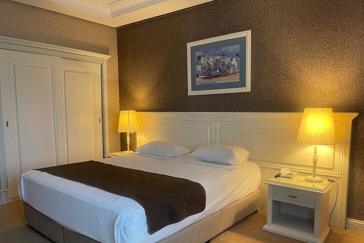 A hotel room with a large bed, two bedside tables with lamps, a painting on the wall, and a door to the left.