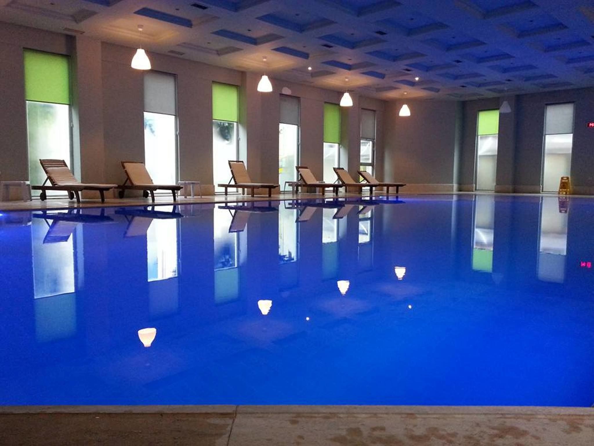 Una piscina interior con agua azul cristalina, múltiples sillas de descanso y grandes ventanales con vidrio teñido de verde.