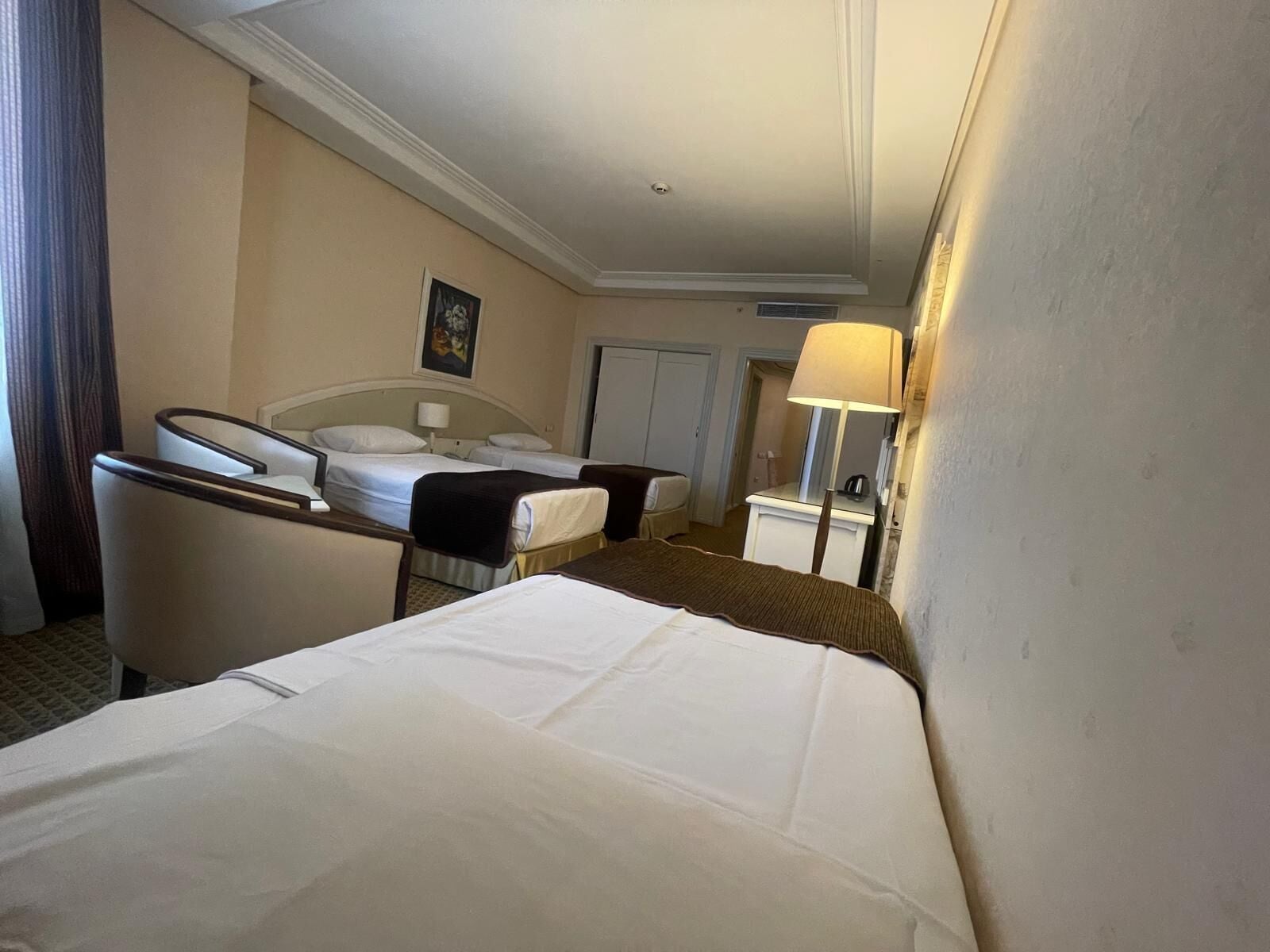 Quarto de hotel com duas camas, uma escrivaninha, uma cadeira, um abajur e um quadro na parede.