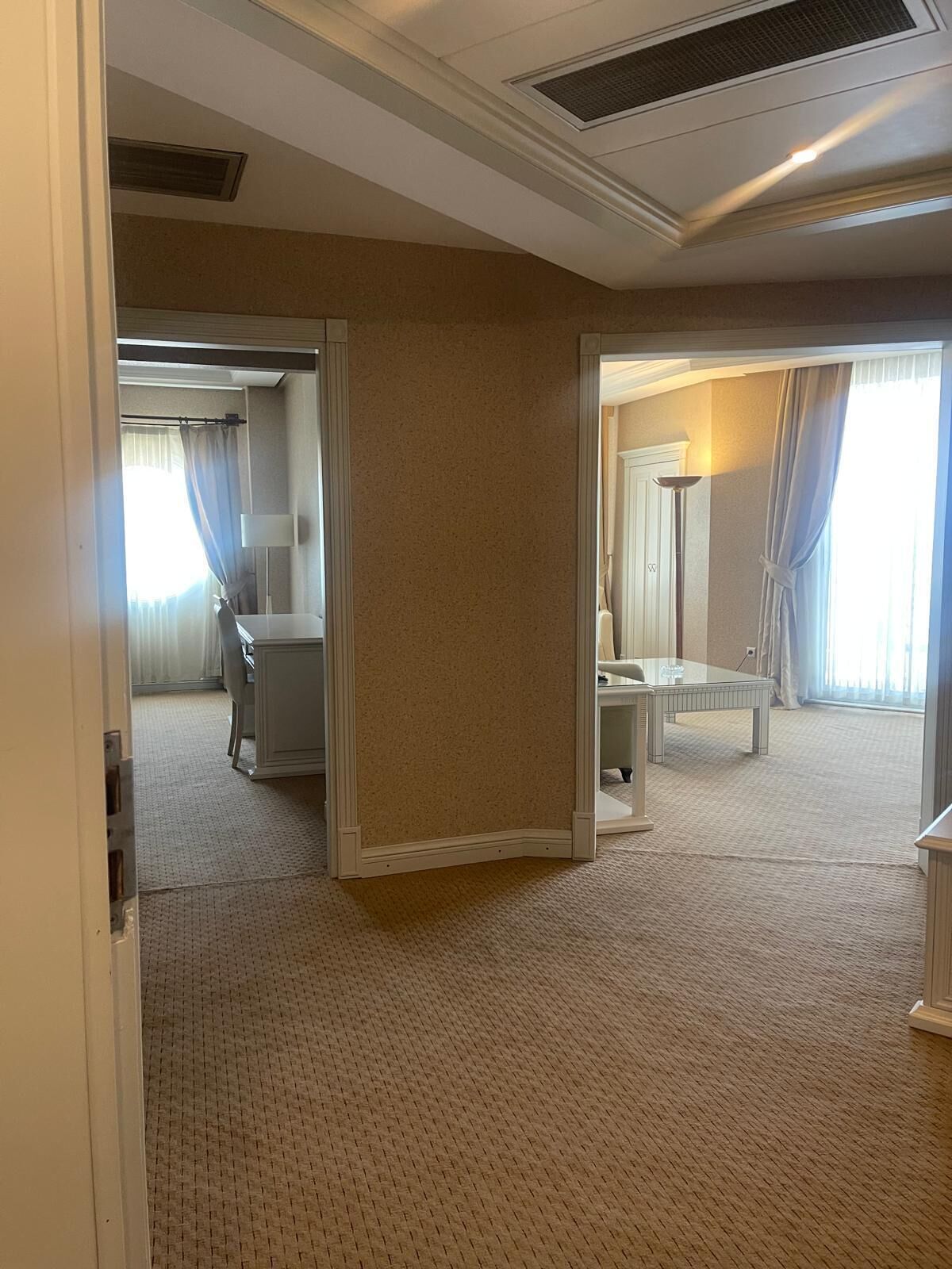 Quarto de hotel com uma cama grande, uma escrivaninha, uma cadeira e uma mesinha. Há uma janela com cortinas e uma luminária na escrivaninha.